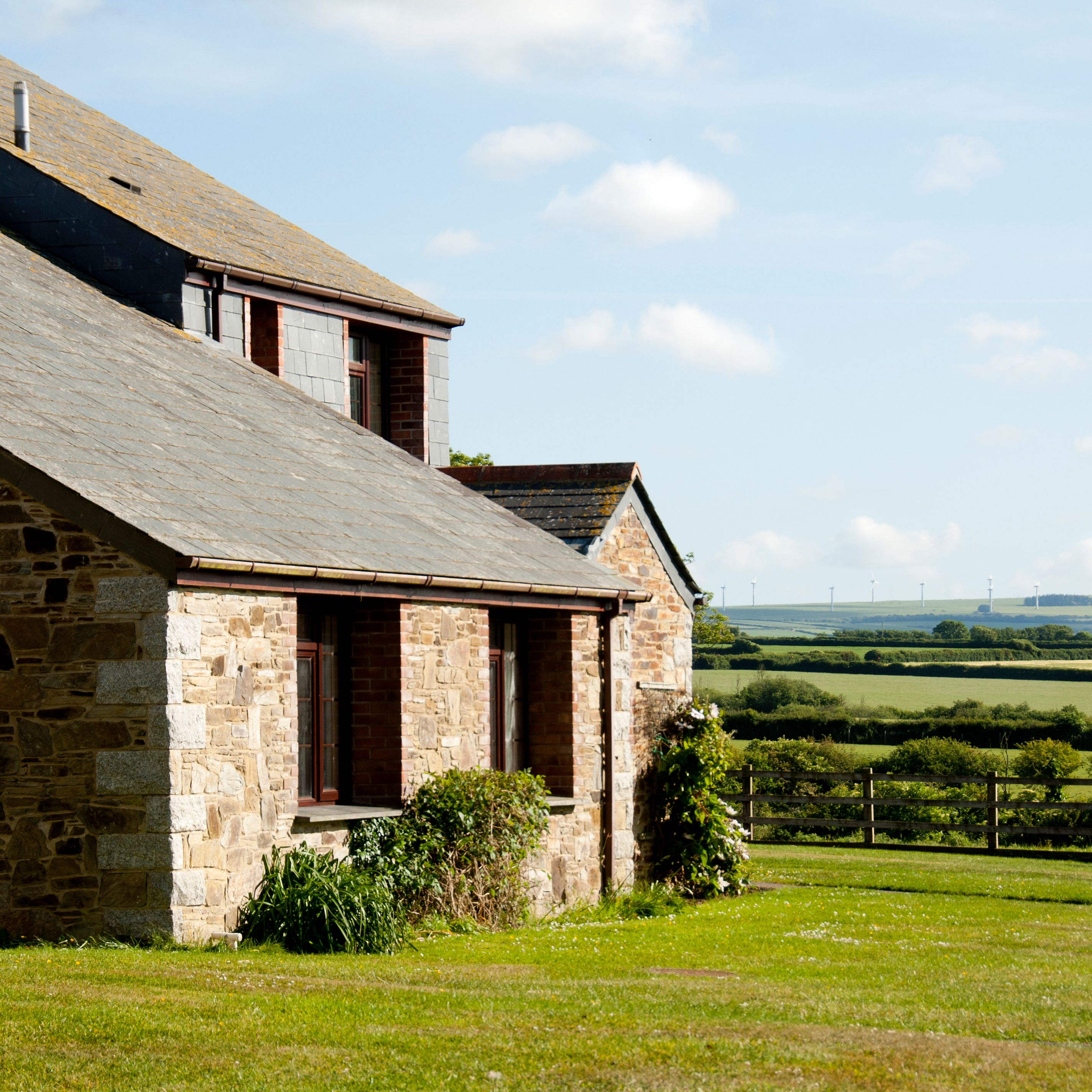 A stone farmhouse with a slate roof in a rural countryside setting, surrounded by green fields under a partly cloudy blue sky.