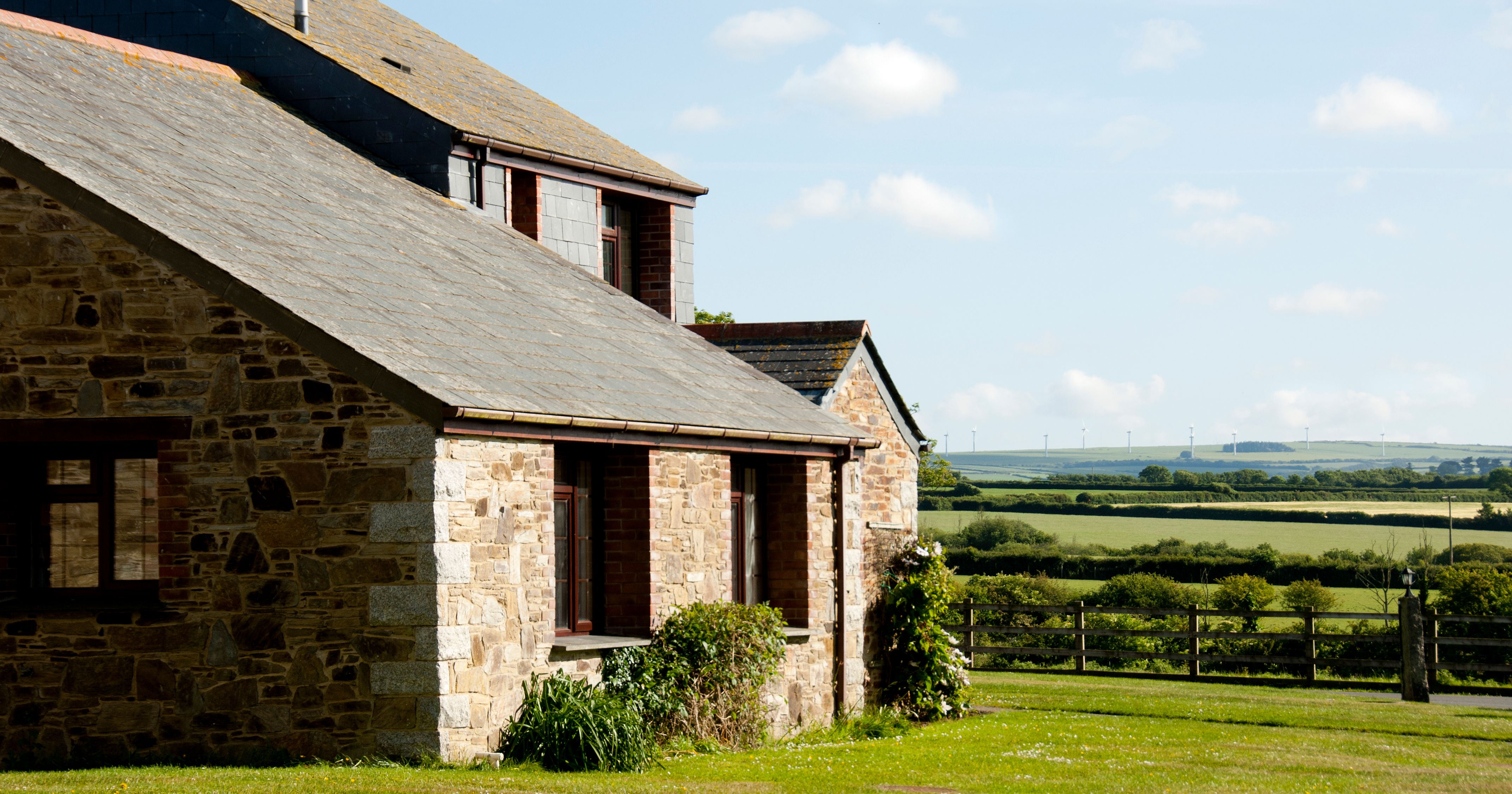 A stone farmhouse with a slate roof in a rural countryside setting, surrounded by green fields under a partly cloudy blue sky.