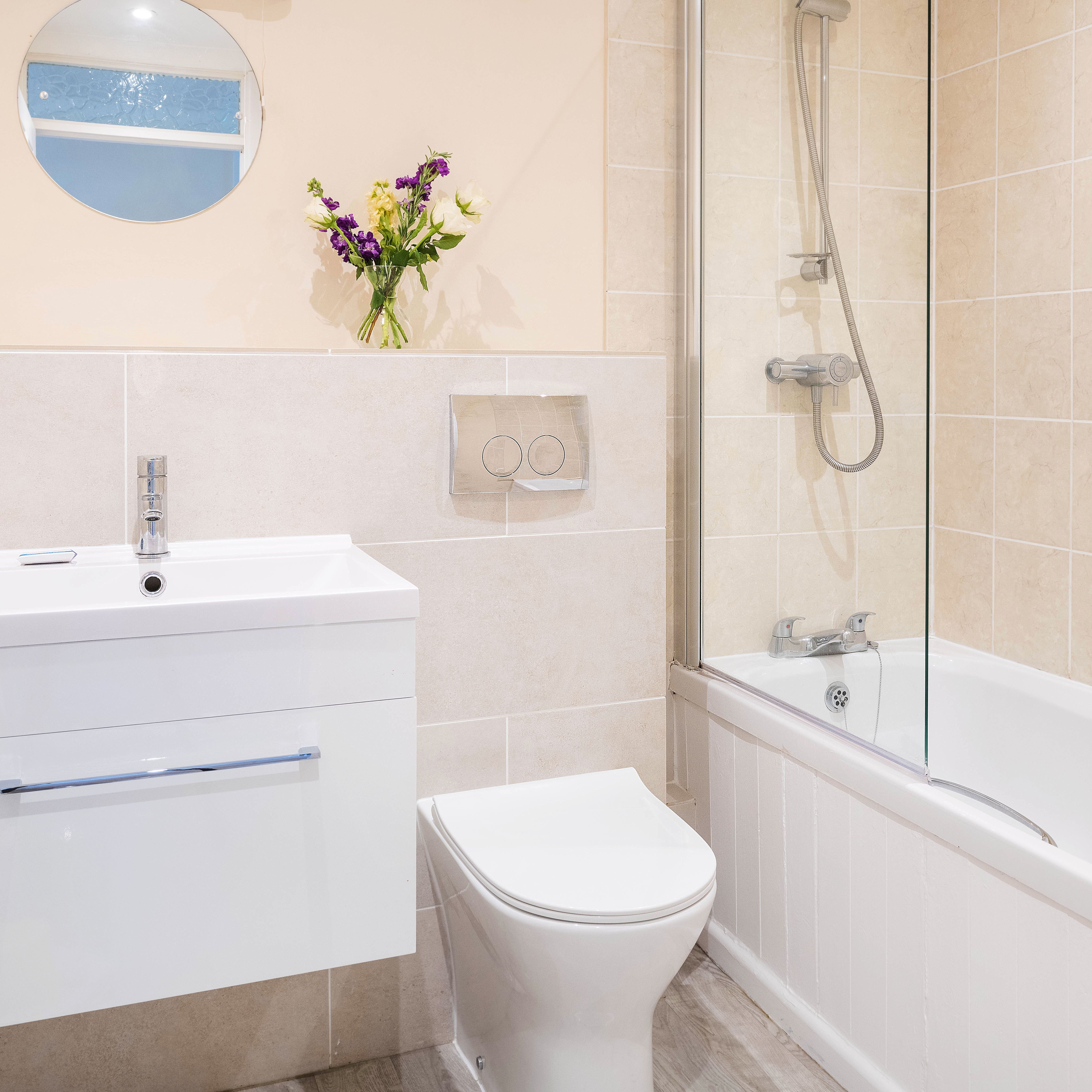 Modern bathroom with a white vanity, toilet, bathtub with glass shower screen, wall-mounted shower, round mirror, vase of flowers, and beige tiled walls.