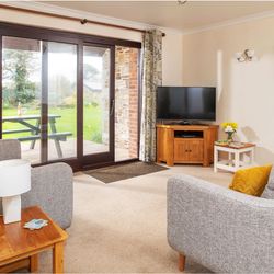 Cozy living room with grey sofas, yellow cushions, wooden furniture, flat screen TV, and sliding glass doors opening to a garden with picnic table.