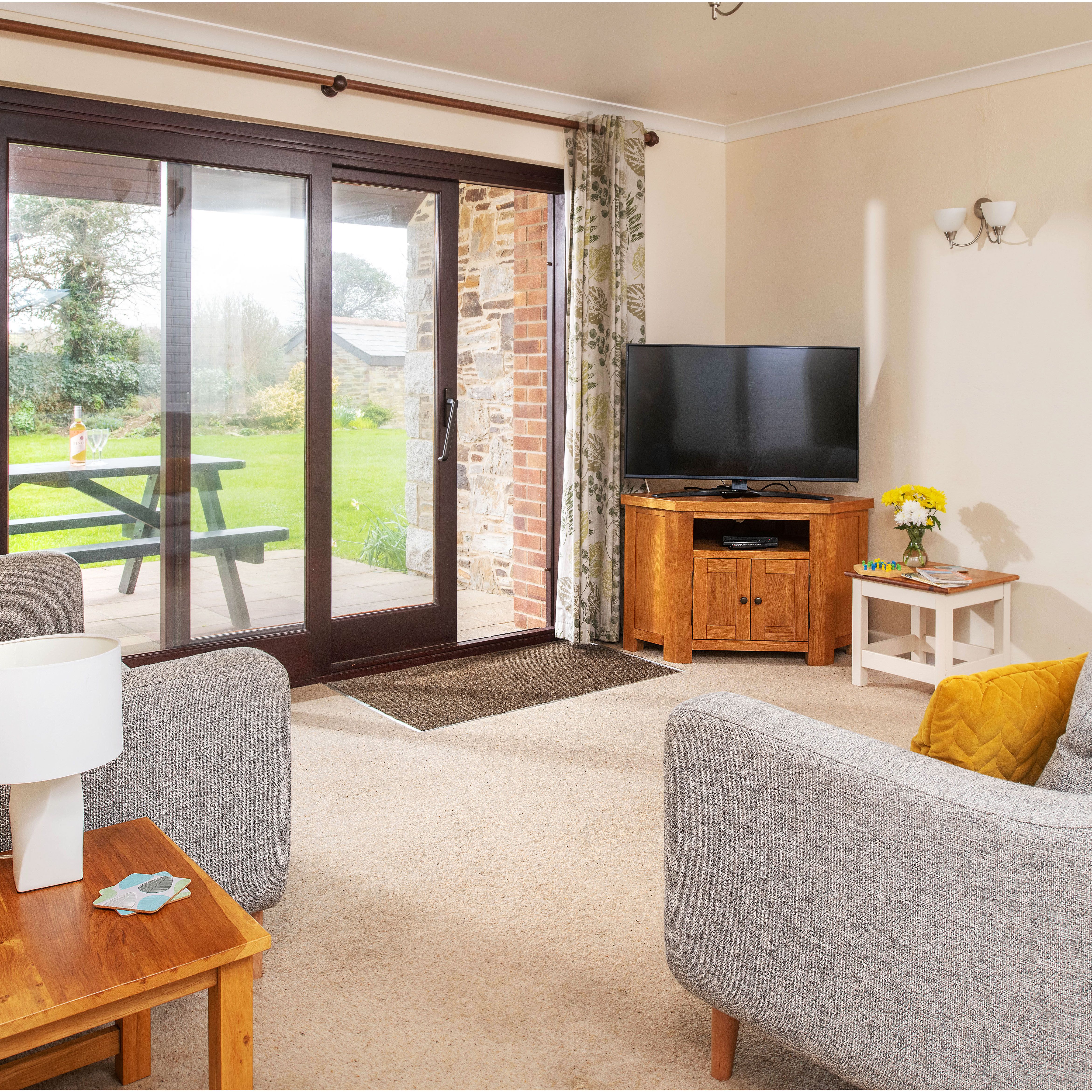 Cozy living room with grey sofas, yellow cushions, wooden furniture, flat screen TV, and sliding glass doors opening to a garden with picnic table.