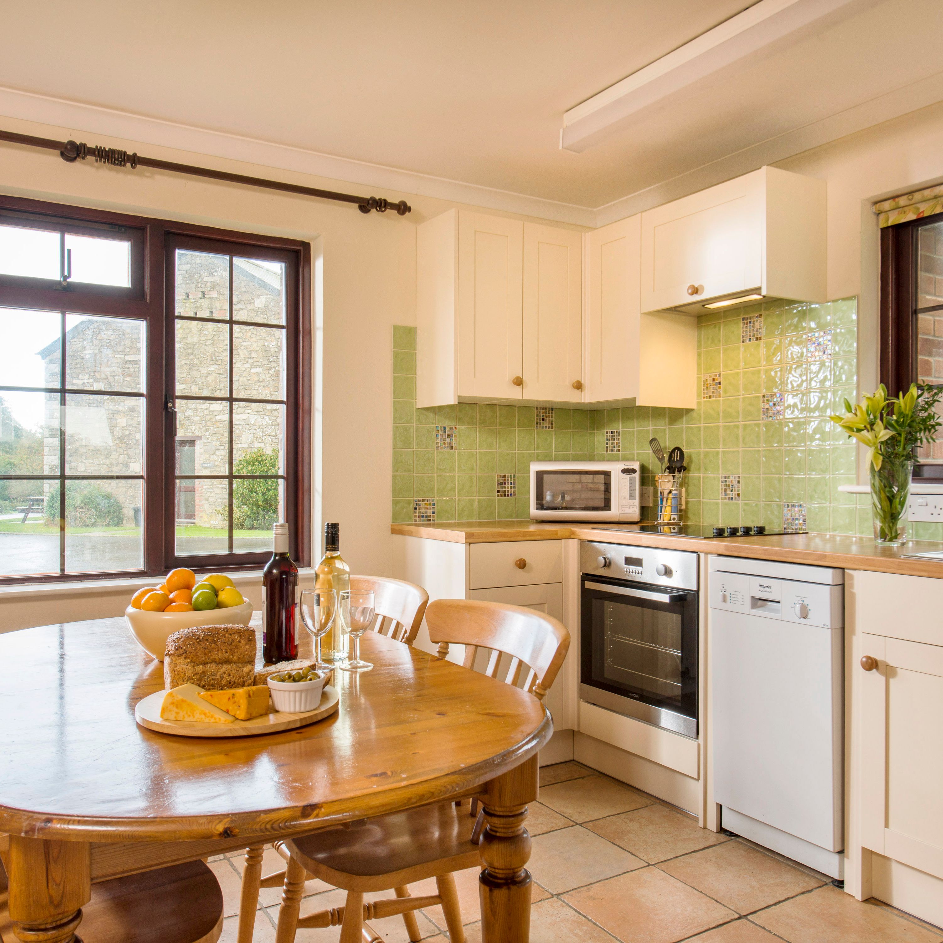 Bright and cozy kitchen with light green tile backsplash, white cabinets, wooden dining table set with food and wine, and large windows overlooking a garden.