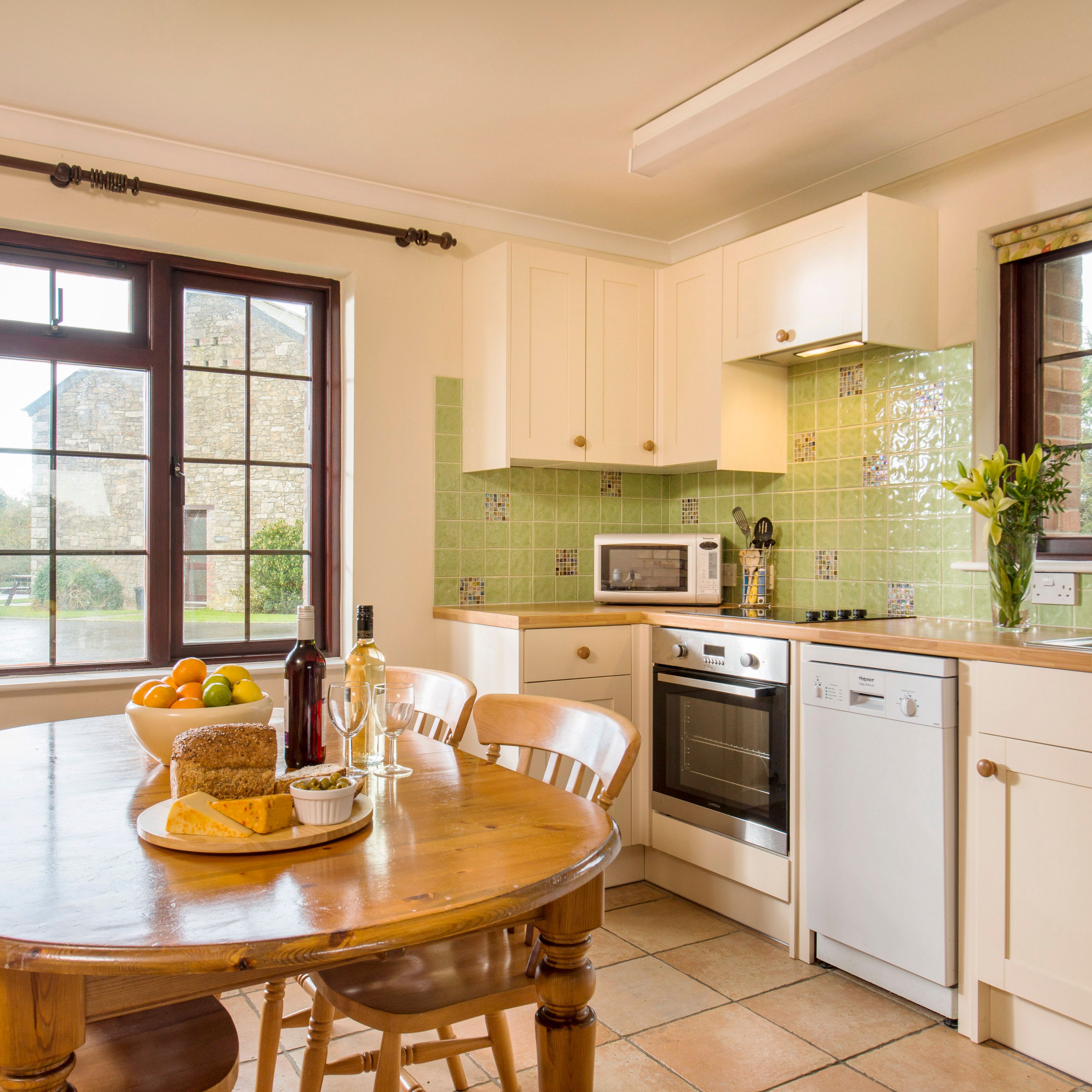 Bright and cozy kitchen with light green tile backsplash, white cabinets, wooden dining table set with food and wine, and large windows overlooking a garden.