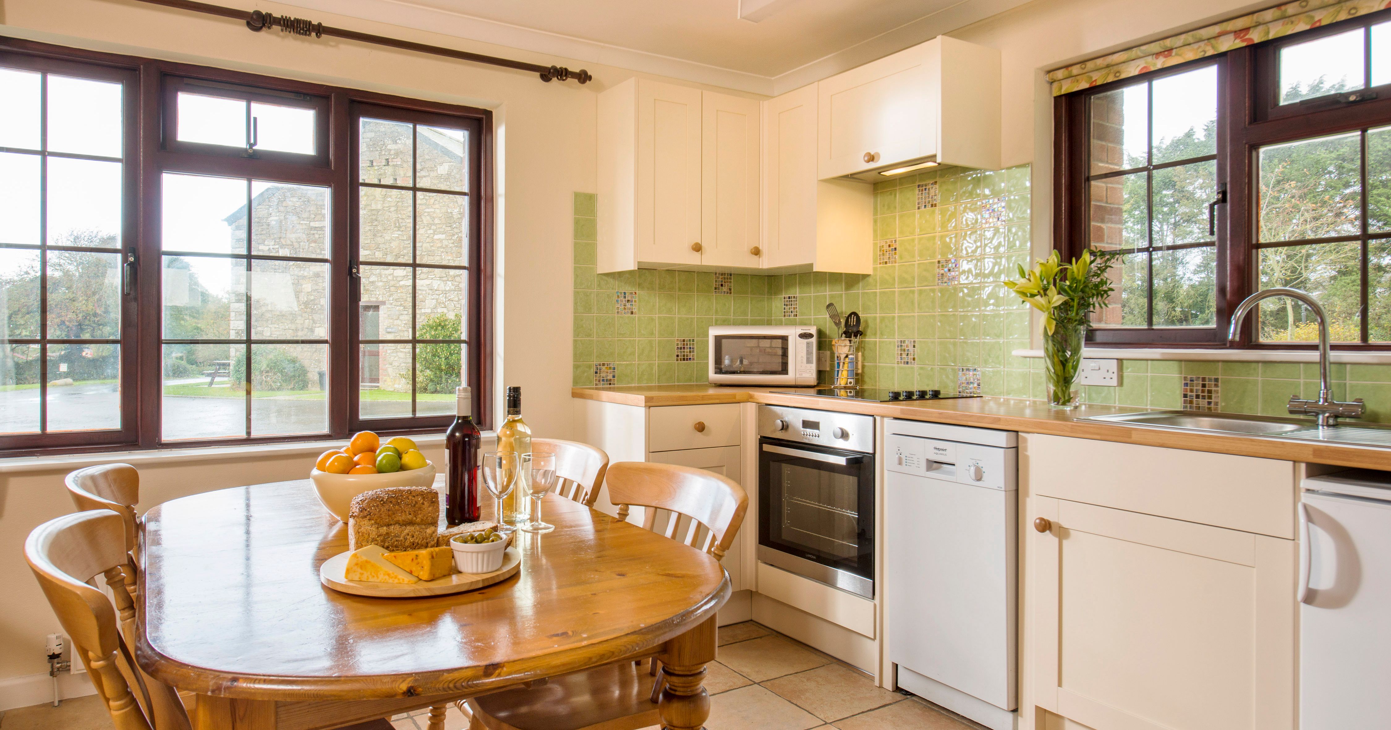 Bright and cozy kitchen with light green tile backsplash, white cabinets, wooden dining table set with food and wine, and large windows overlooking a garden.