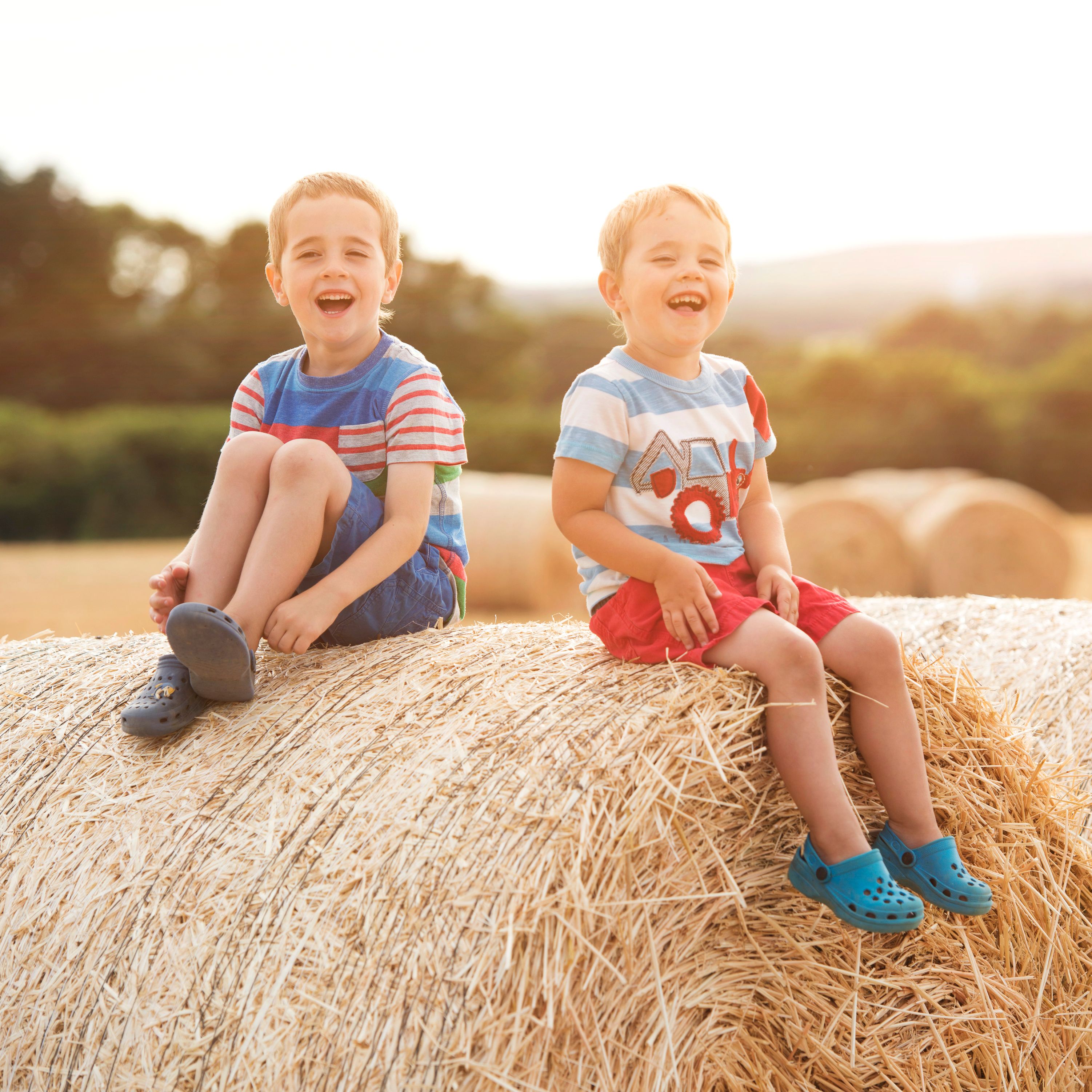 Two young boys sitting and laughing on a large round hay bale in a sunny field with more hay bales in the background.