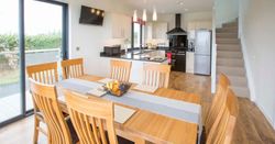 Modern dining area with wooden table and six chairs, a kitchen with white cabinets and black appliances, a basket of fruit on the table, sliding glass doors, and a staircase leading upstairs.