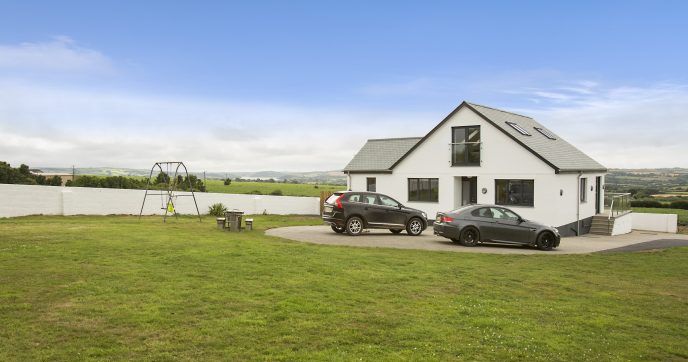 Modern two-story white house with grey roof, two cars parked in the driveway, large green lawn, outdoor swing set, and distant view of rolling hills under a blue sky.