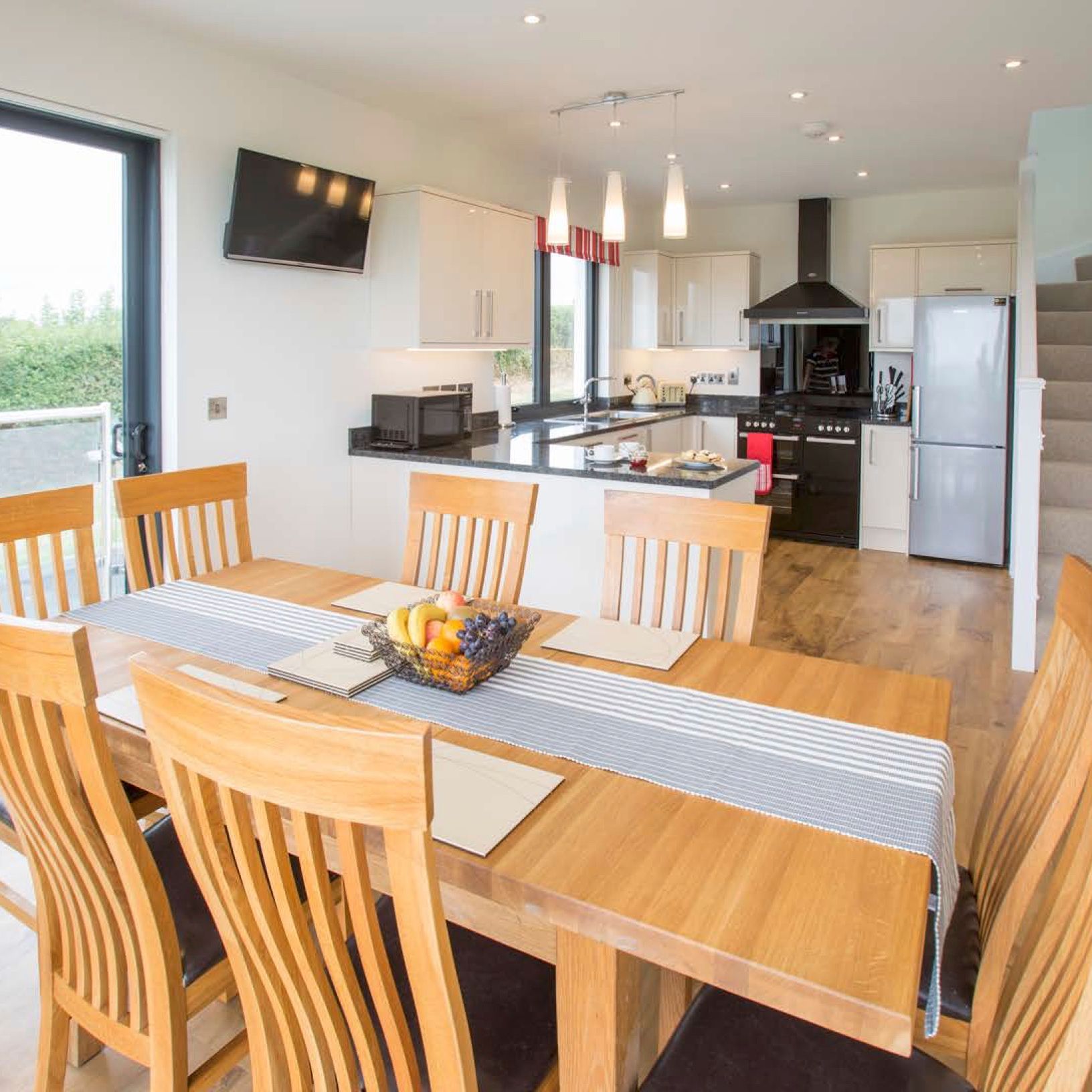 Modern open-plan kitchen and dining area with wooden table and chairs, a fruit basket on the table, sliding glass doors, and a staircase leading upstairs.