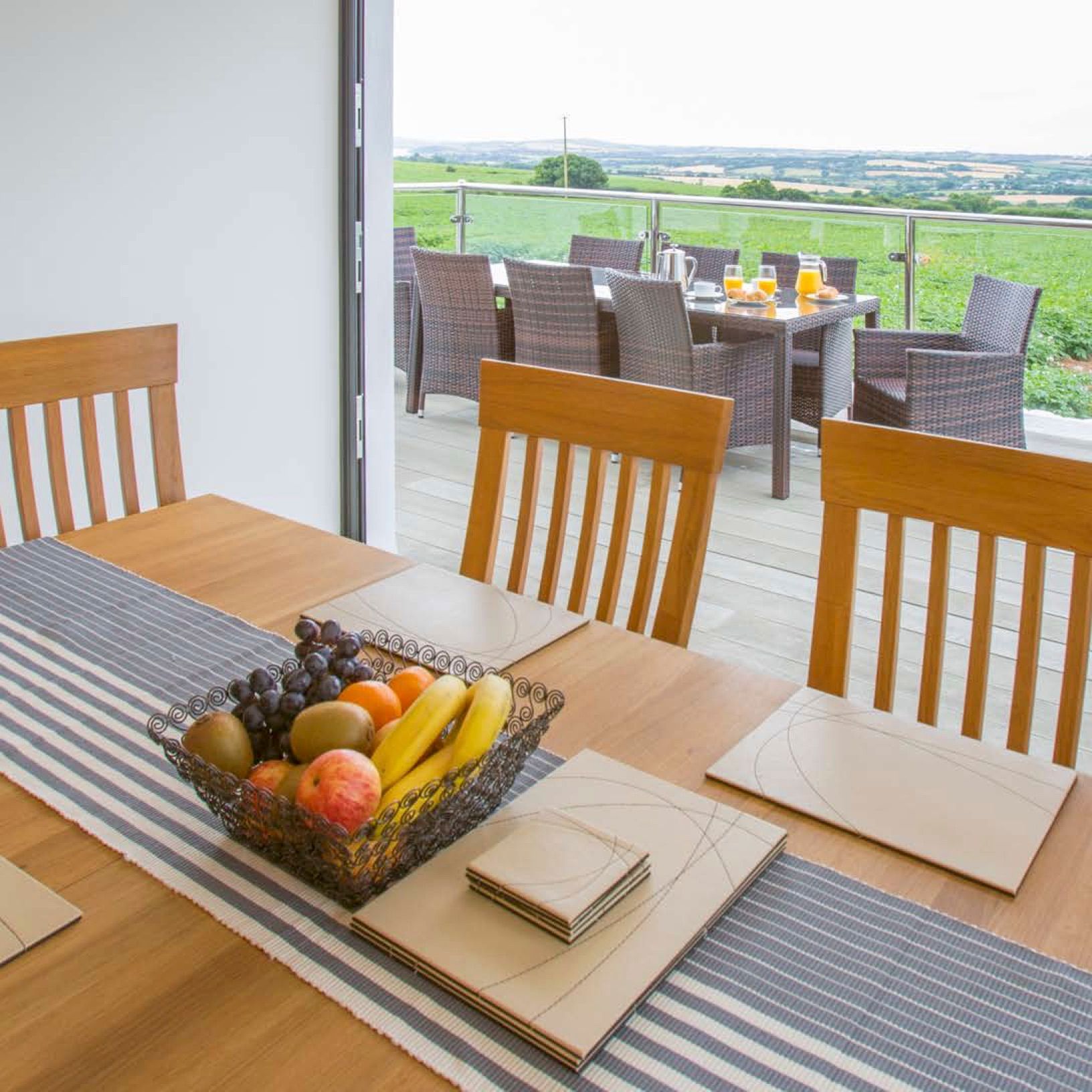 A modern dining table set with placemats and a fruit basket inside a house, with a view of an outdoor patio set for breakfast and a scenic countryside landscape in the background.