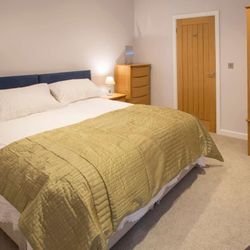 Modern bedroom with a large bed featuring white bedding and a green quilt, wooden wardrobe and dresser, bedside lamp, and a wooden door.