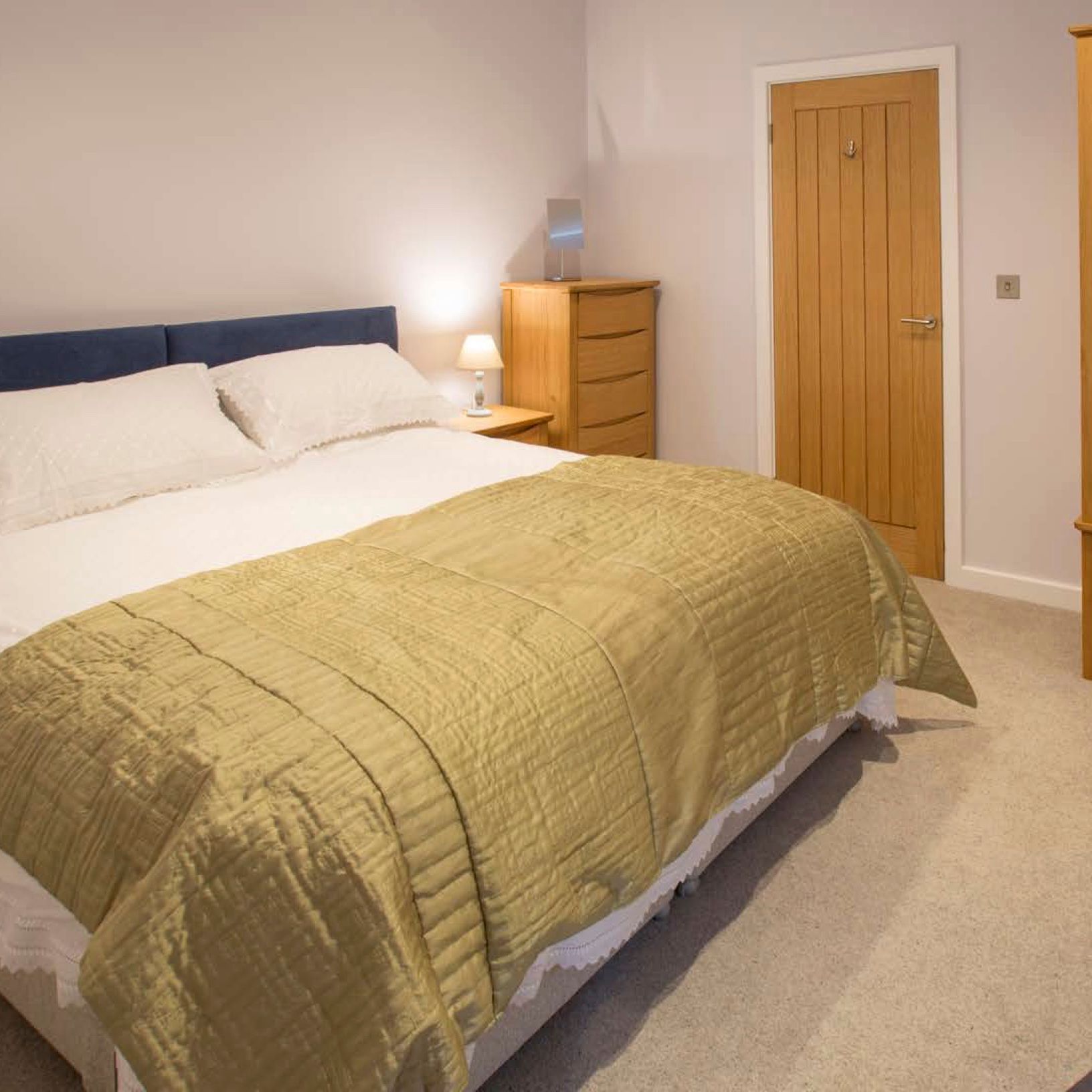 Modern bedroom with a large bed featuring white bedding and a green quilt, wooden wardrobe and dresser, bedside lamp, and a wooden door.