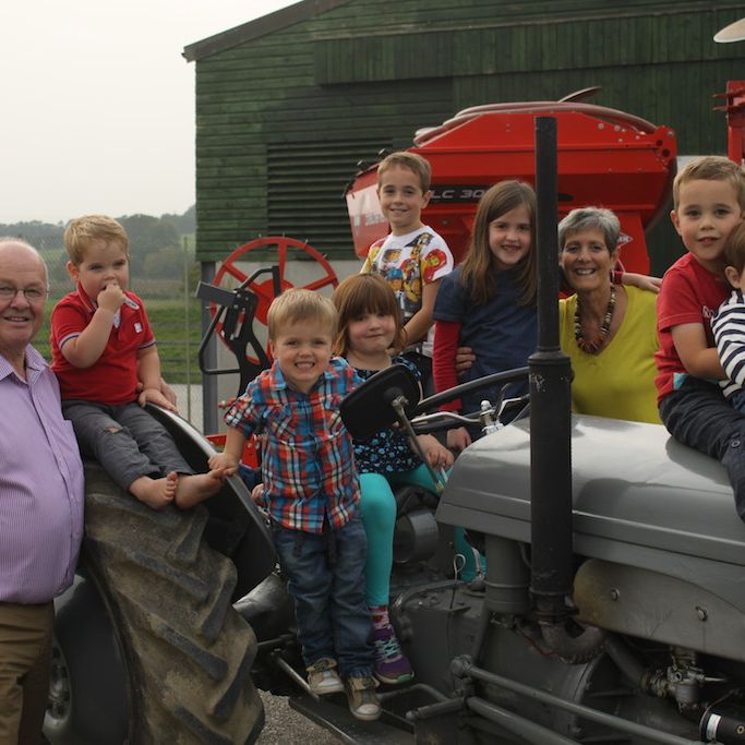 A group of children and two adults sitting and standing on and around a vintage tractor, smiling for the camera in an outdoor farm setting with another tractor and barn in the background.