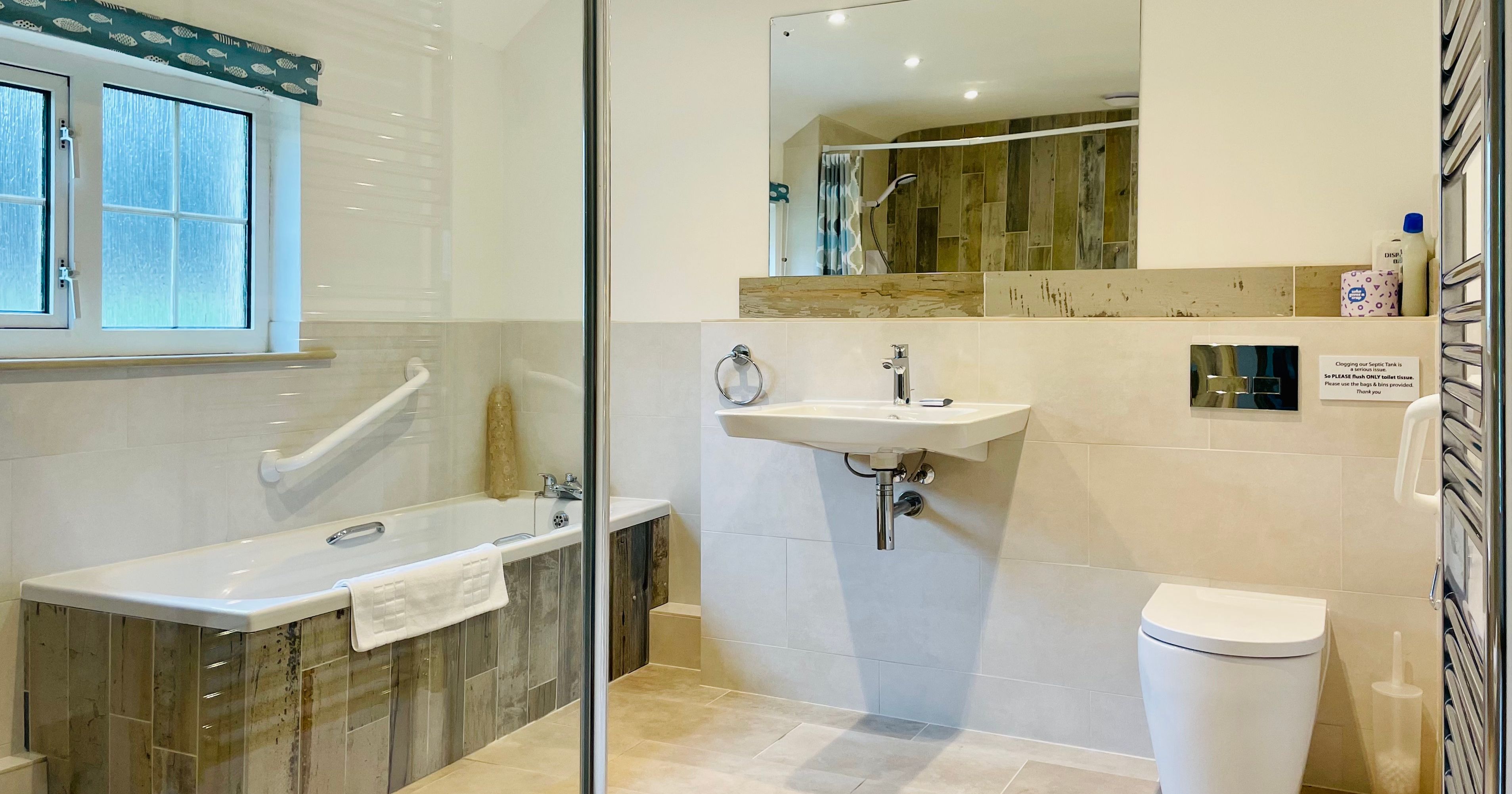 Modern accessible bathroom with bathtub, grab bars, wall-mounted sink, toilet, large mirror, window with patterned blind, and beige tiled flooring and walls.