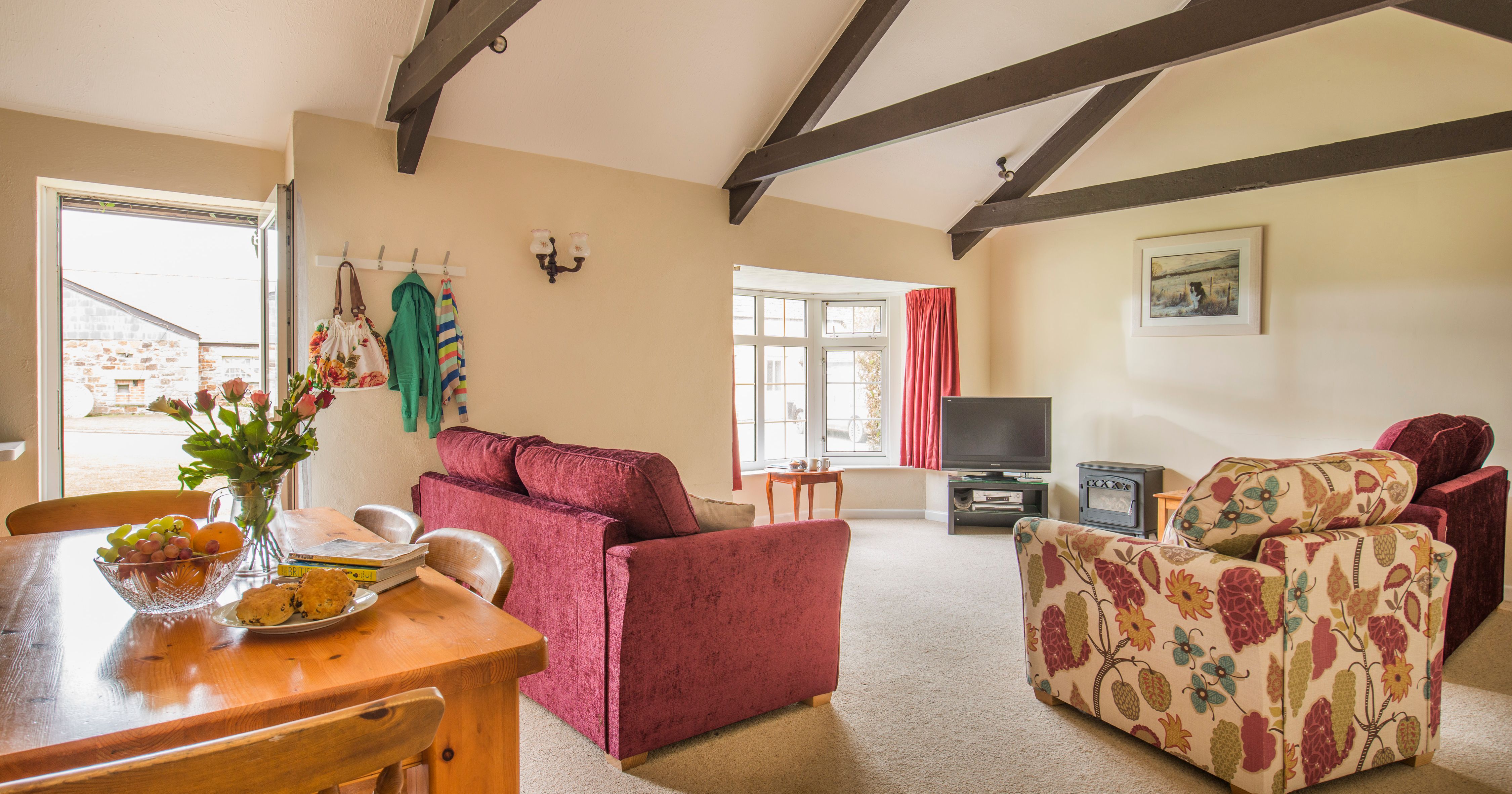 Cozy living room with exposed wooden beams, red velvet sofas, a floral armchair, dining table with flowers and fruit, TV, and large window with red curtains.