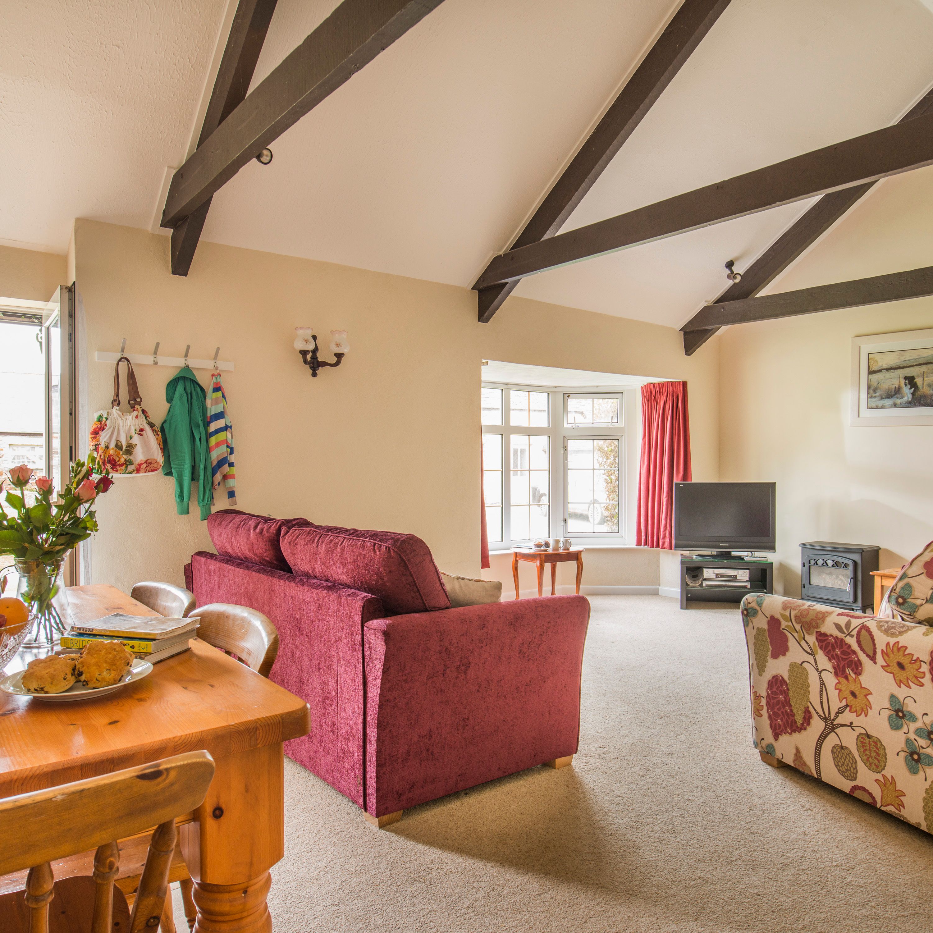 Cozy living room with exposed wooden beams, red velvet sofas, a floral armchair, dining table with flowers and fruit, TV, and large window with red curtains.