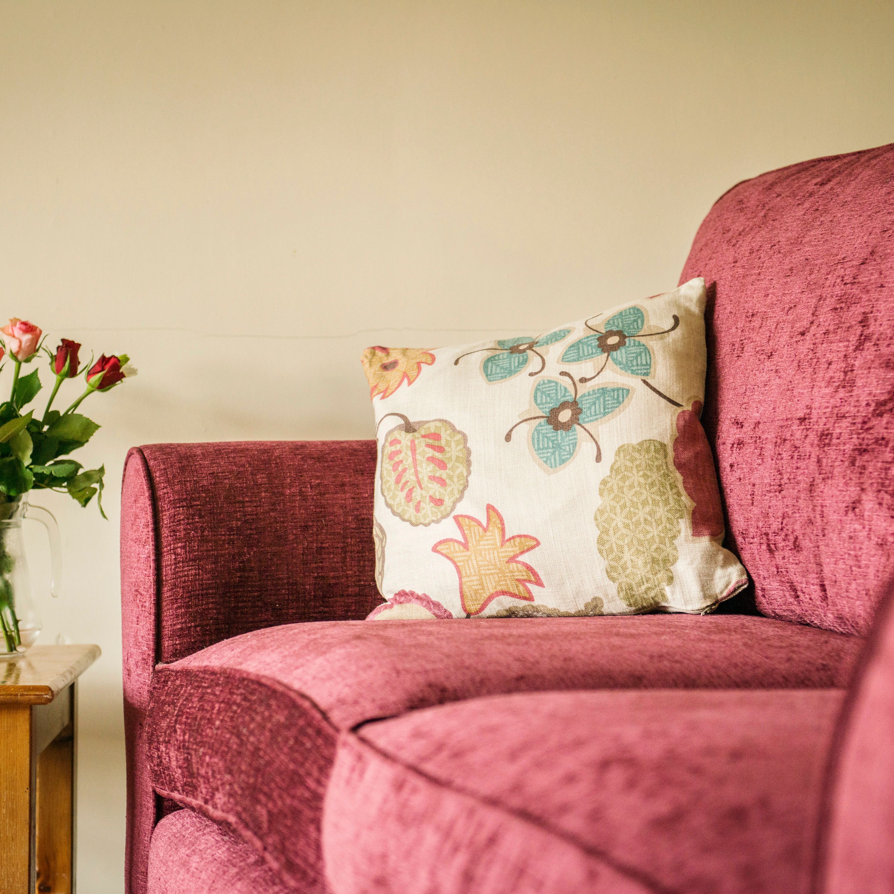A cozy pink sofa with a floral cushion, next to a wooden side table holding a vase of pink and red roses.