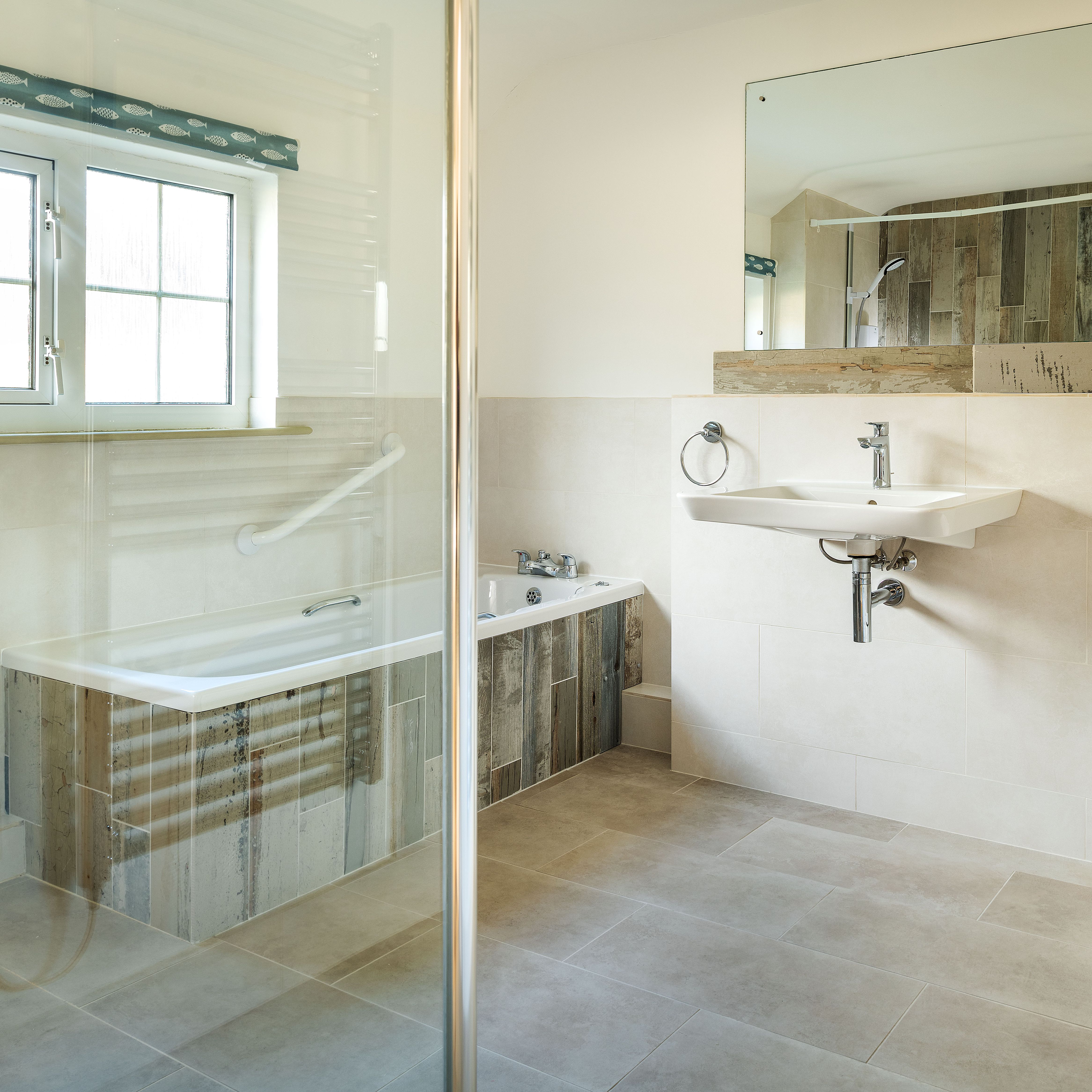 Modern bathroom with a bathtub, white sink, toilet, glass shower enclosure, large wall mirror, beige tiled floor and walls, and a vase with flowers on a shelf.