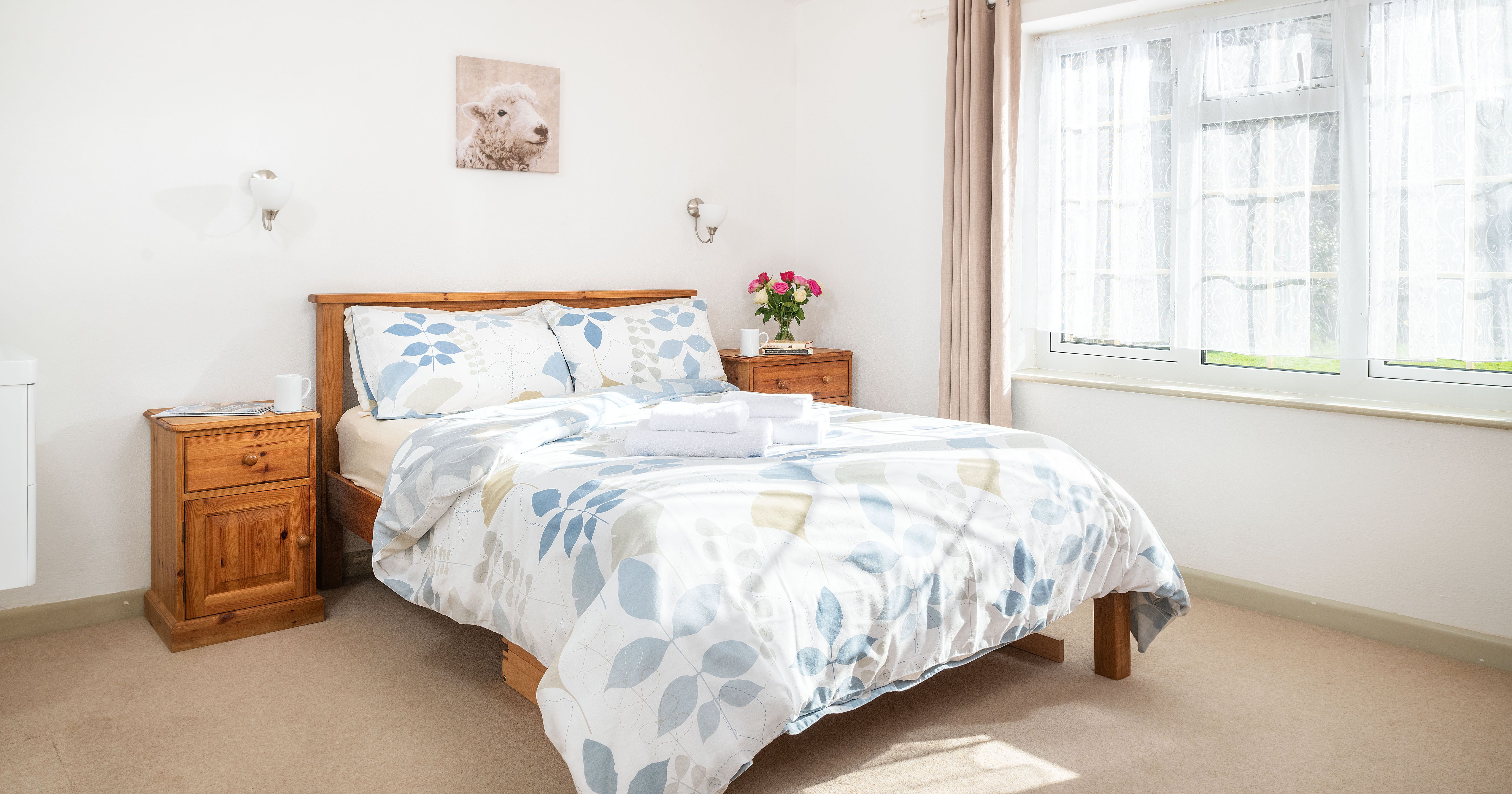Bright and cozy bedroom with wooden bed frame, floral bedding, two wooden nightstands, flowers, and a large window with sheer curtains.