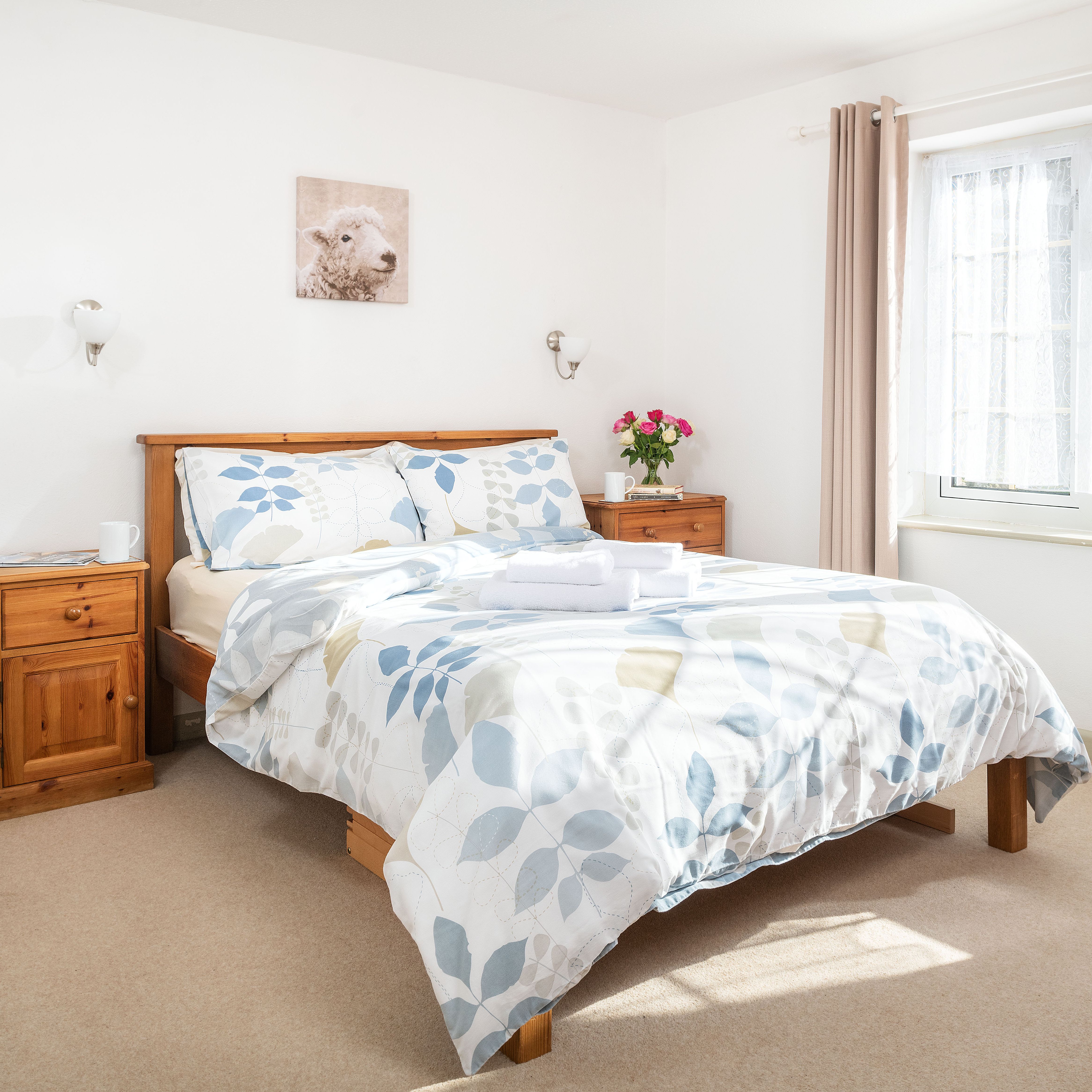 Bright and cozy bedroom with wooden bed frame, floral bedding, two wooden nightstands, flowers, and a large window with sheer curtains.