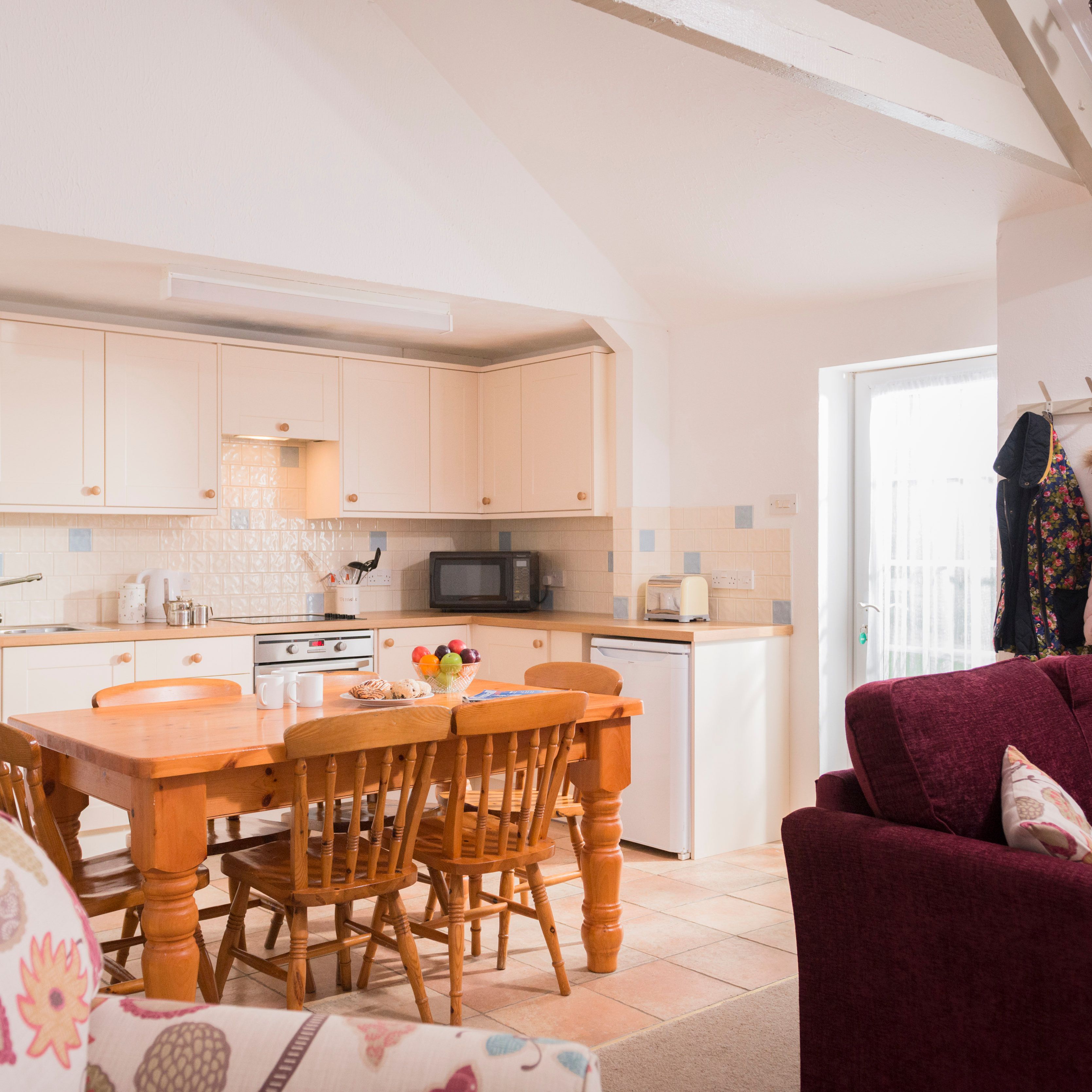 Bright and cozy kitchen and dining area with wooden table and chairs, cream cabinets, a sofa with patterned cushions, and a coat rack with jackets and a pink coat hanging by the door.