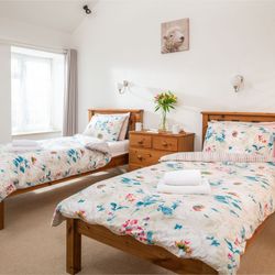 Bright bedroom with two single wooden beds, floral bedding, a wooden dresser with flowers, and a window with light curtains.