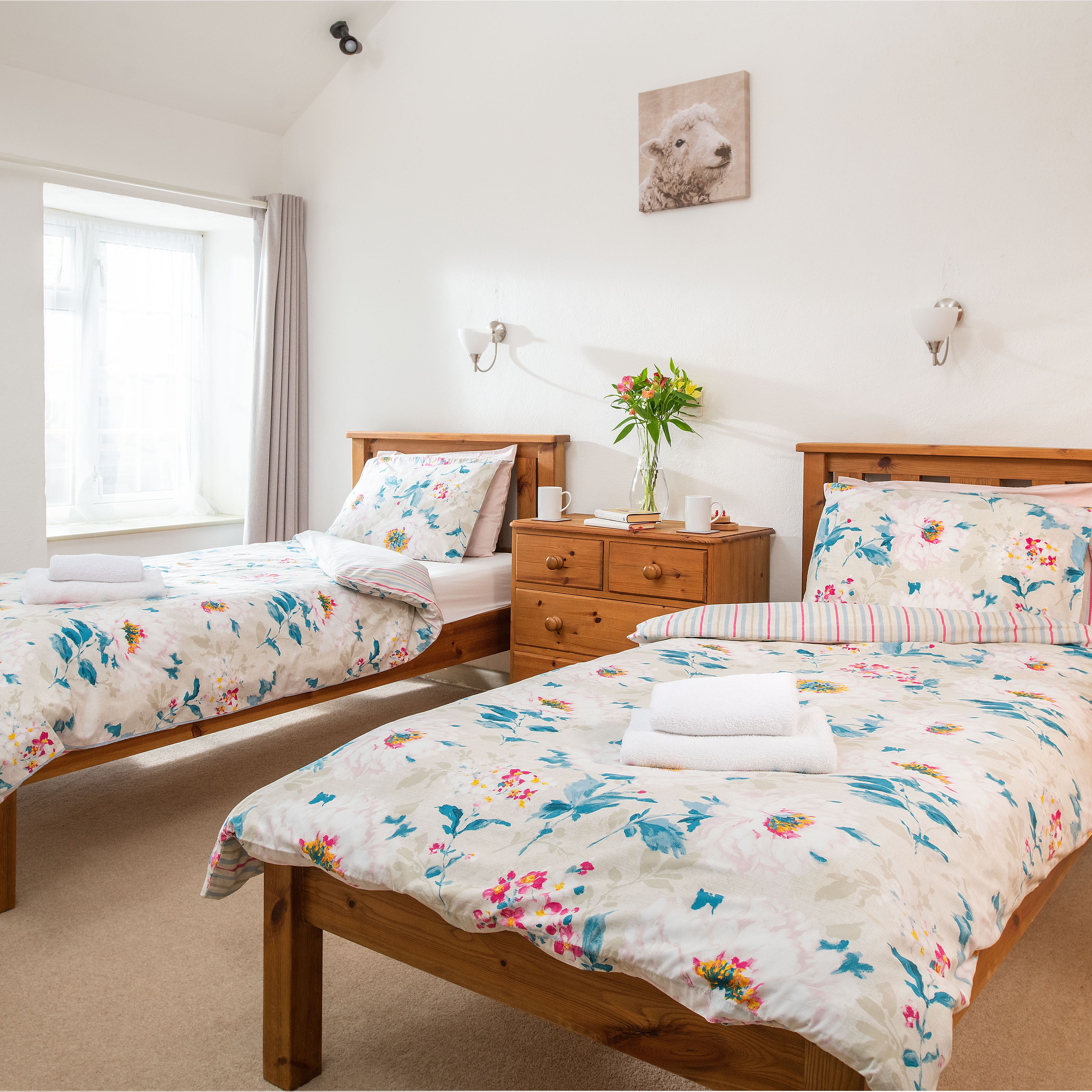 Bright bedroom with two single wooden beds, floral bedding, a wooden dresser with flowers, and a window with light curtains.