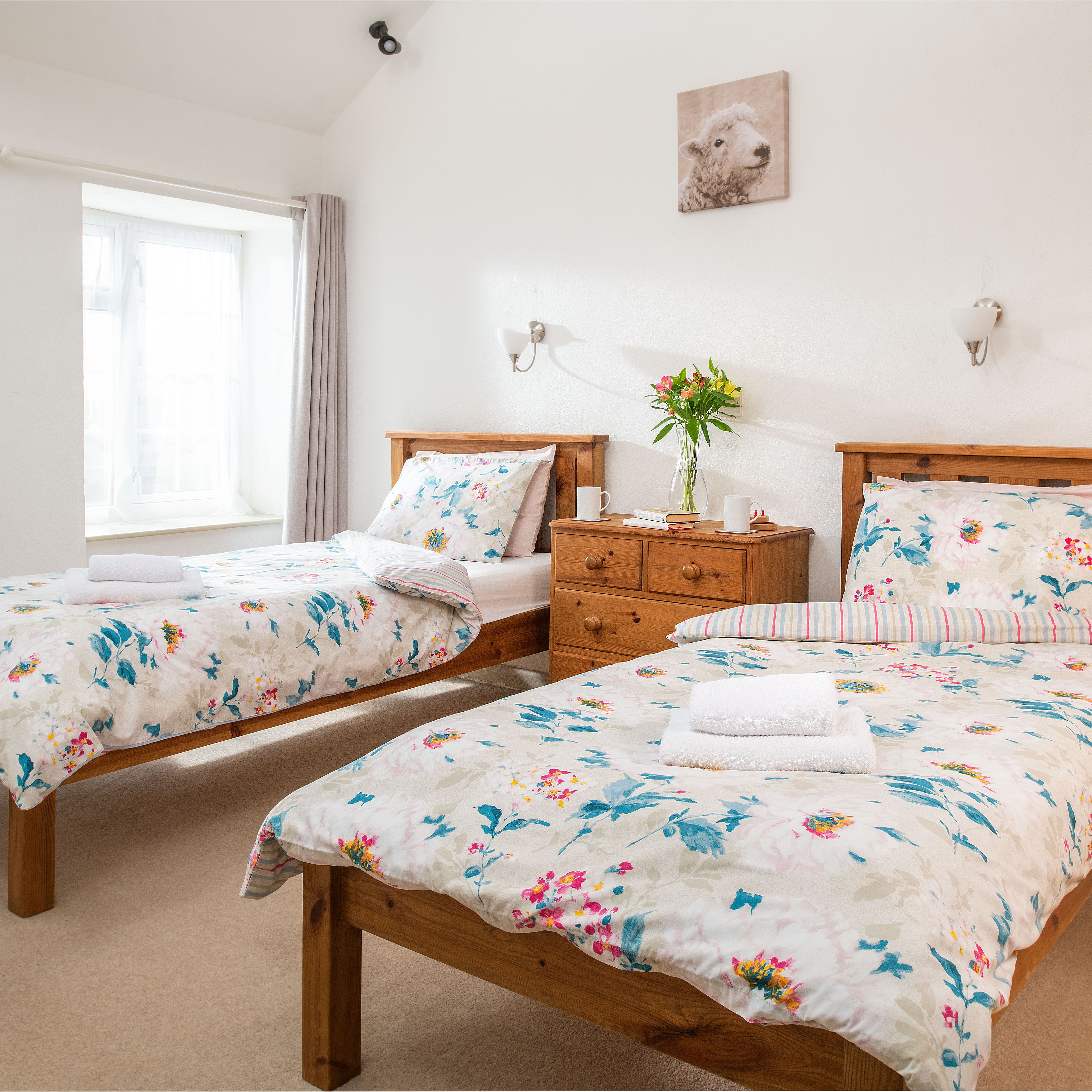 Bright bedroom with two single wooden beds, floral bedding, a wooden dresser with flowers, and a window with light curtains.