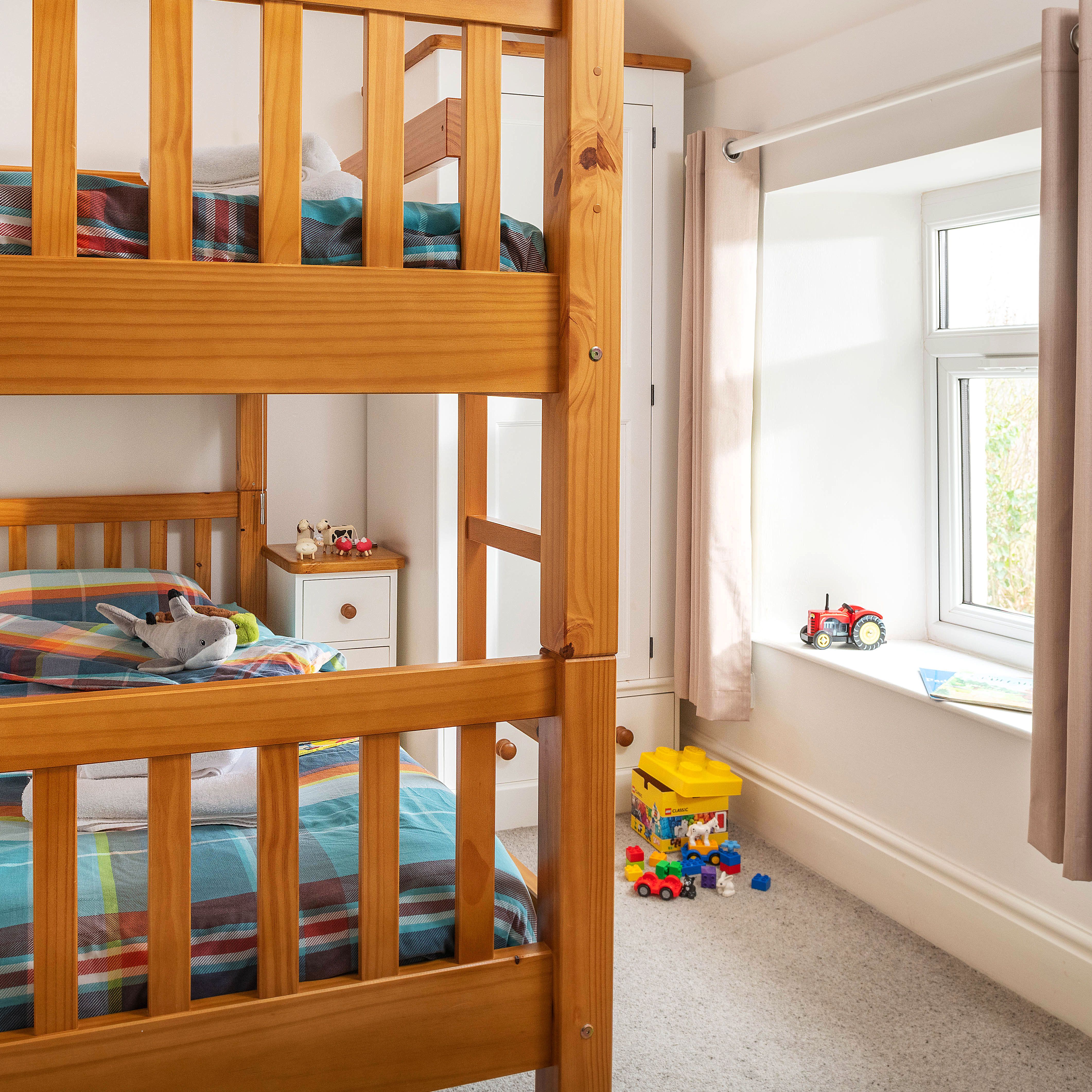 A cozy children's bedroom with wooden bunk beds, plaid bedding, toys on the floor, a window with beige curtains, and a bedside table with small decorations.