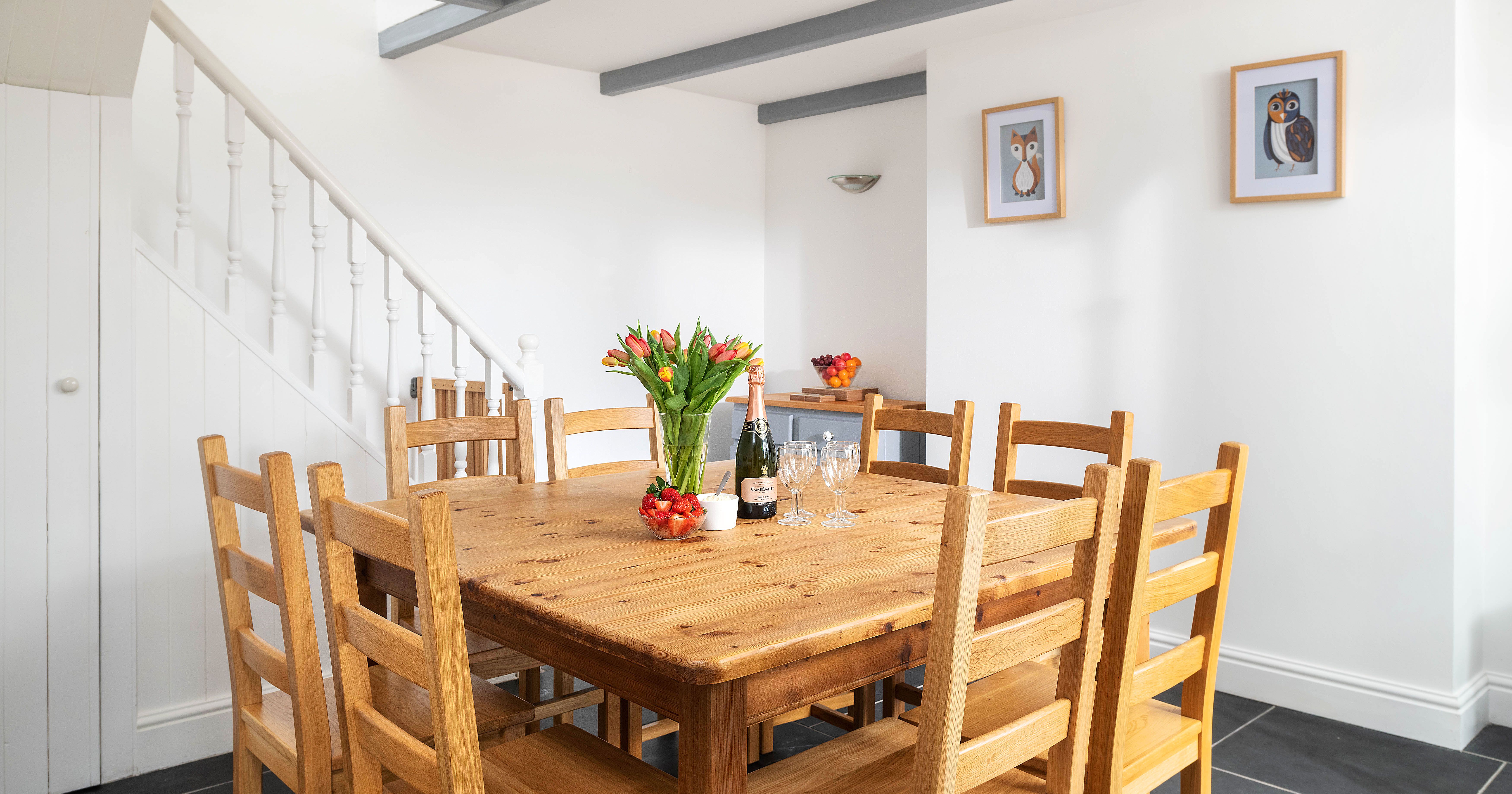 A bright dining room with a wooden table and eight chairs, a vase of tulips, a bottle of wine, strawberries, wine glasses, and framed owl artwork on the white walls.