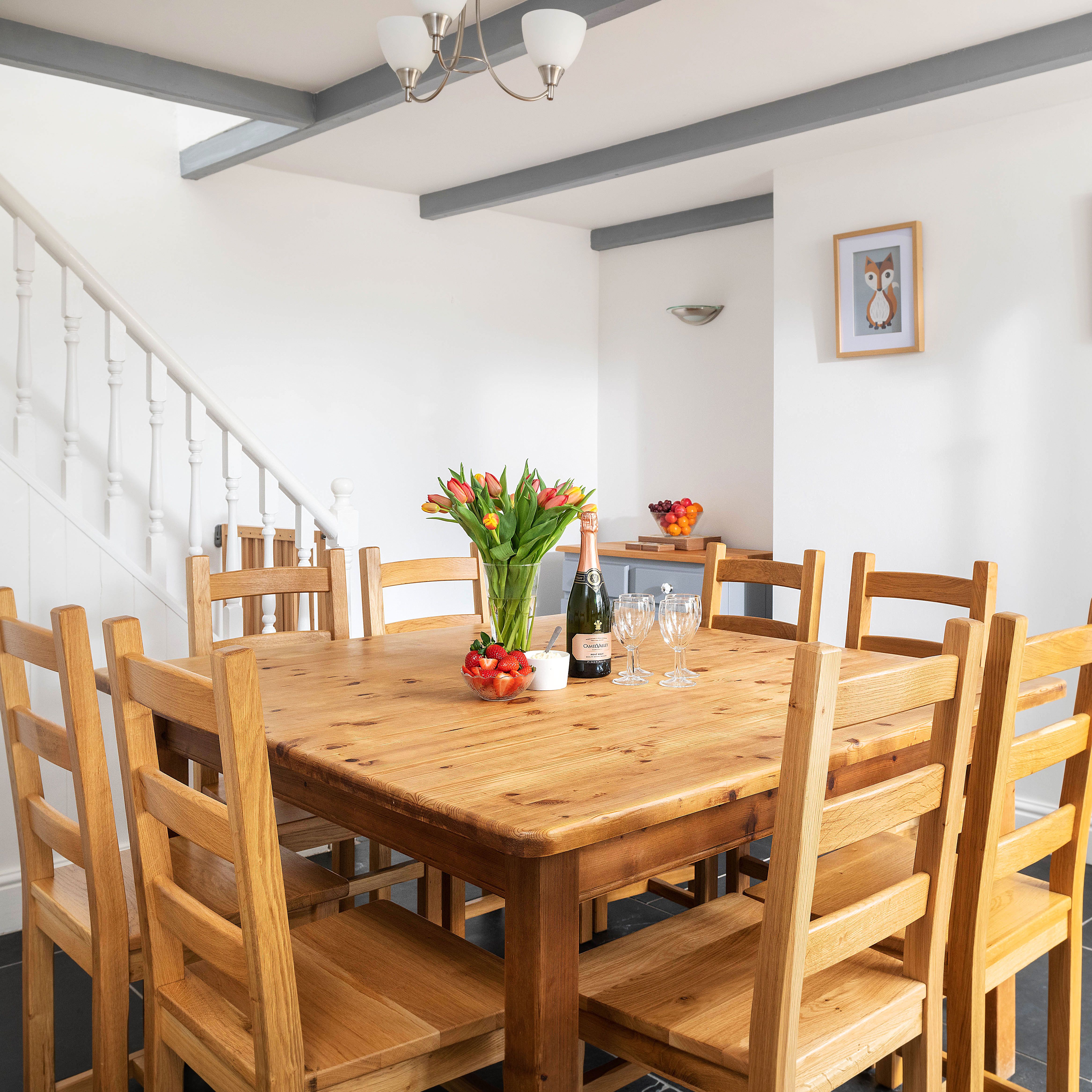 A bright dining room with a wooden table and eight chairs, a vase of tulips, a bottle of wine, strawberries, wine glasses, and framed owl artwork on the white walls.