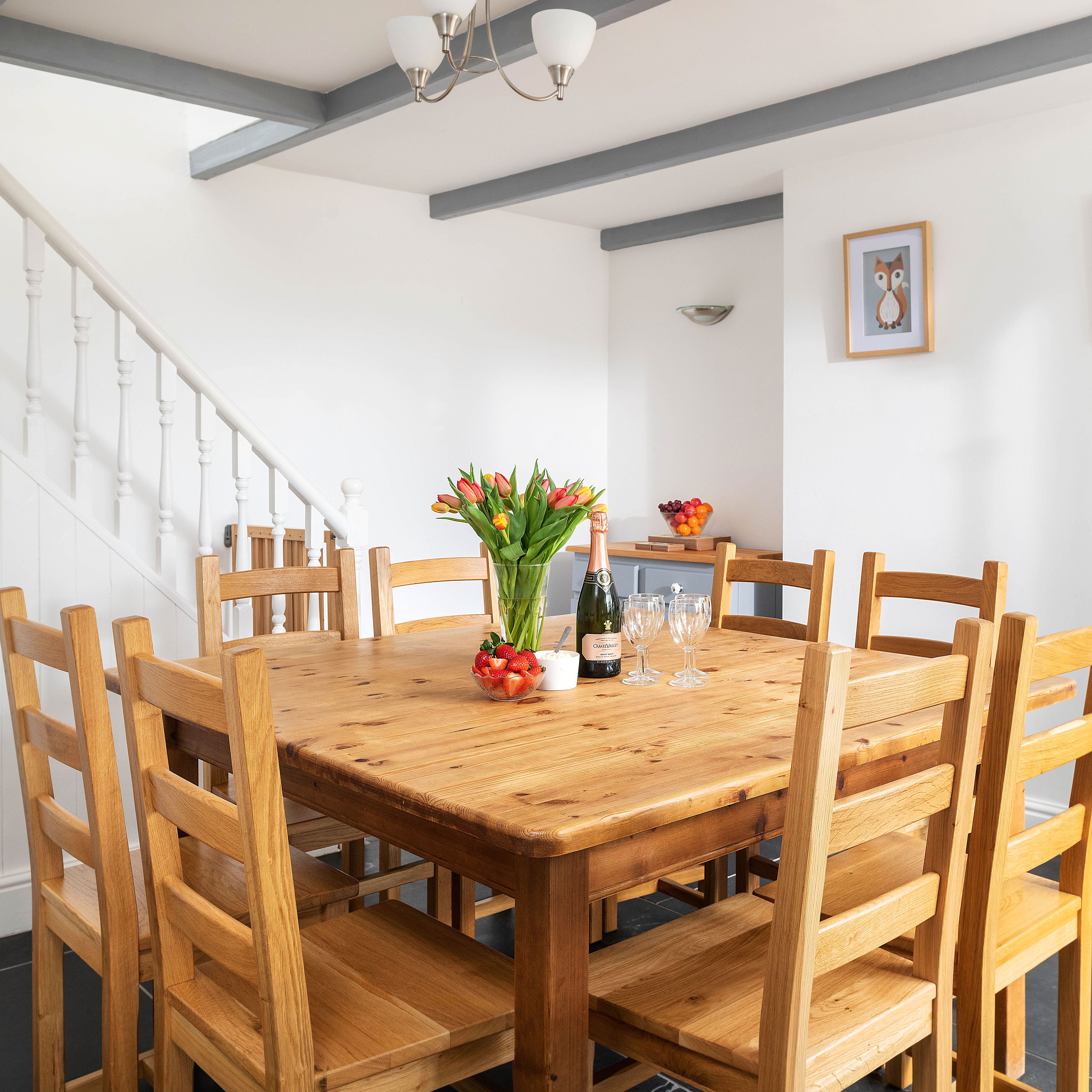 A bright dining room with a wooden table and eight chairs, a vase of tulips, a bottle of wine, strawberries, wine glasses, and framed owl artwork on the white walls.