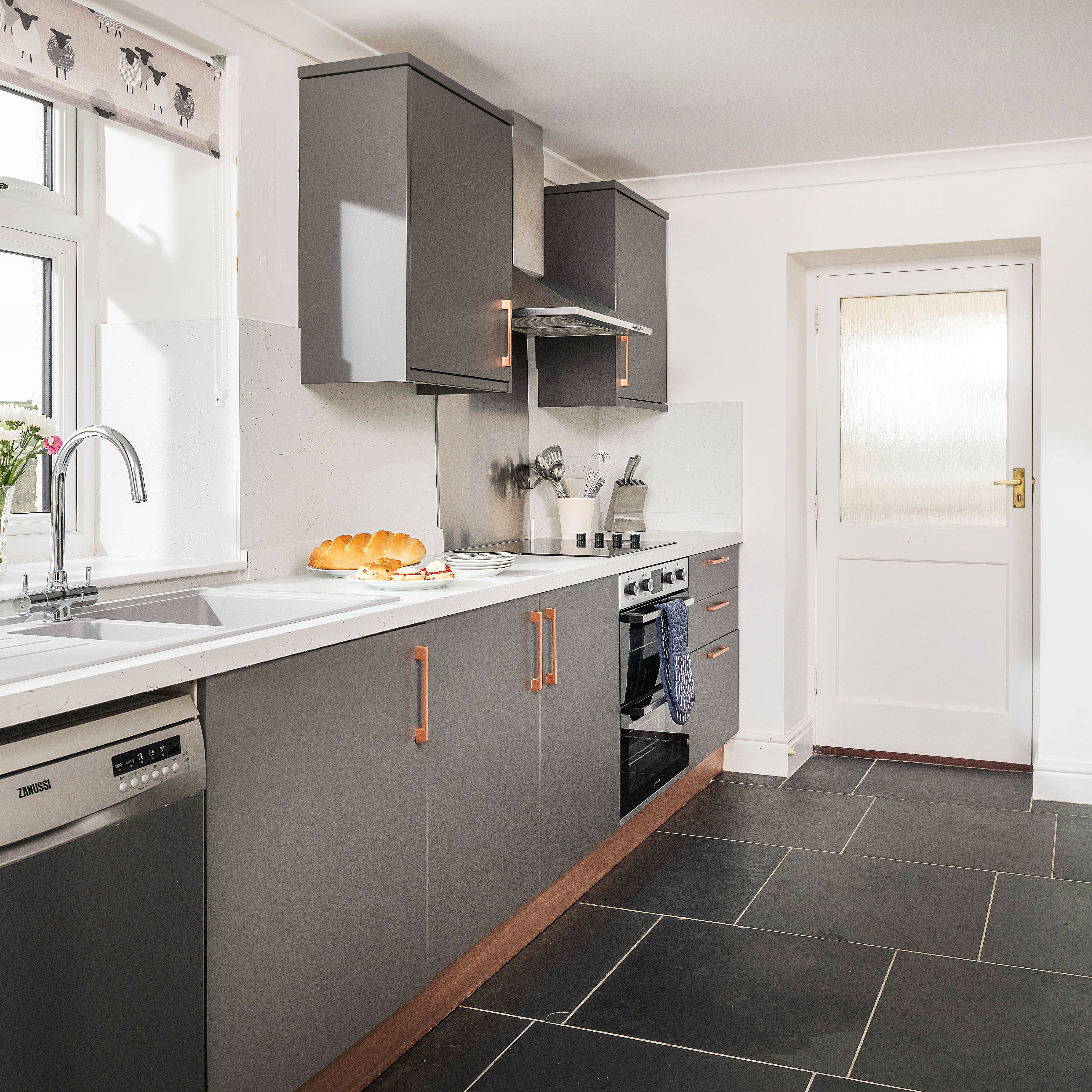 Modern kitchen with grey cabinets, white countertops, stainless steel appliances, and a bowl of fruit and flowers near the sink.