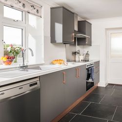 Modern kitchen with grey cabinets, white countertops, stainless steel appliances, and a bowl of fruit and flowers near the sink.
