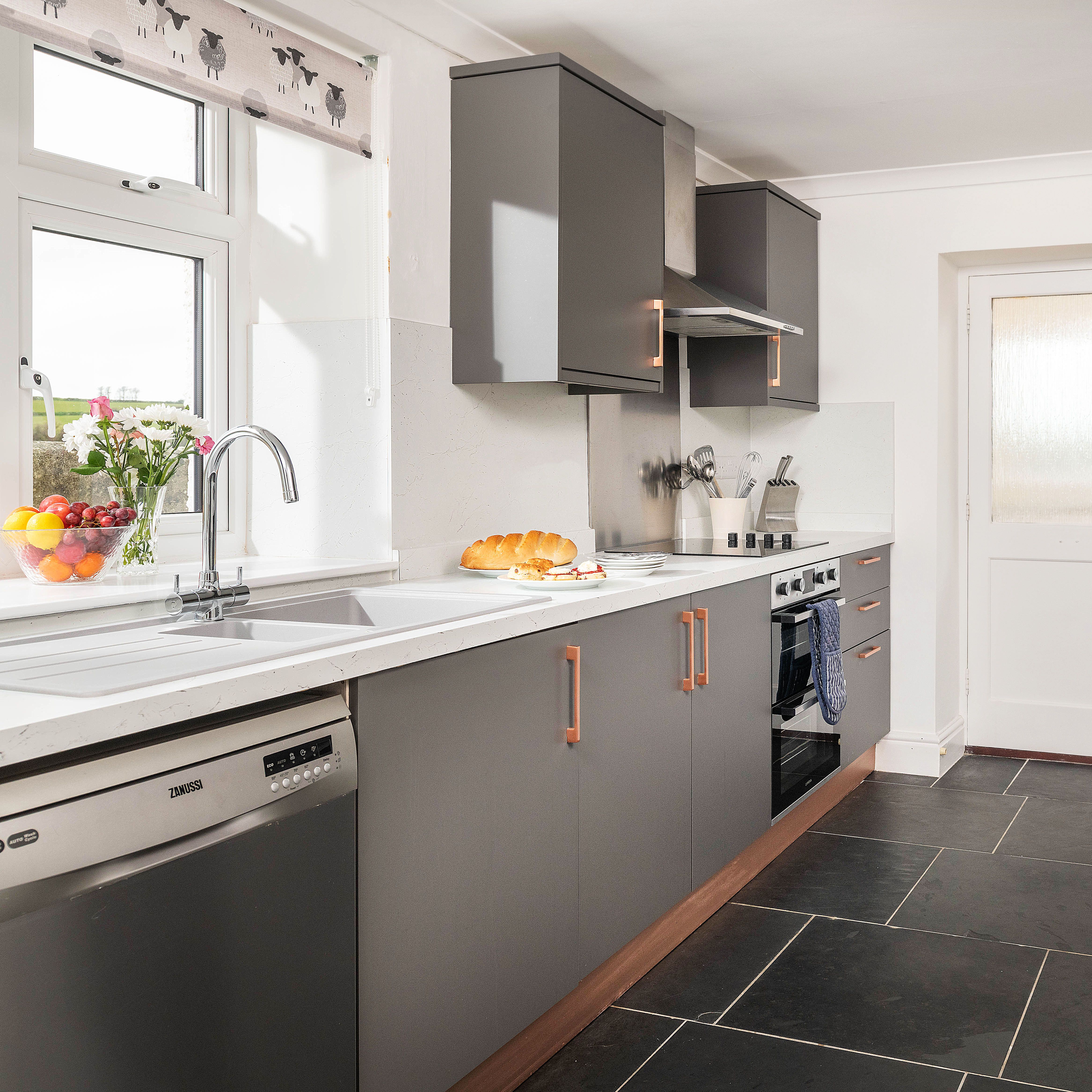 Modern kitchen with grey cabinets, white countertops, stainless steel appliances, and a bowl of fruit and flowers near the sink.