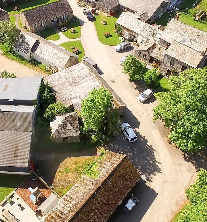 Aerial view of a rural village with several stone buildings, green lawns, trees, roads, and parked cars.