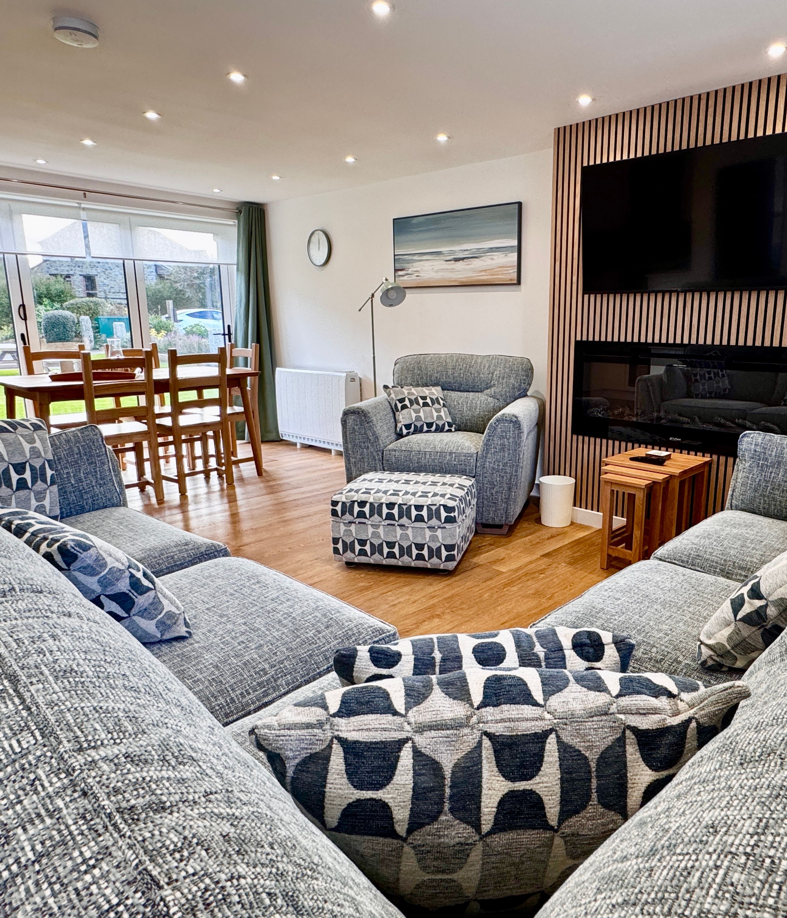 Modern living room with gray sectional sofa and patterned cushions, wooden flooring, wall-mounted TV above a fireplace, dining table by large windows with garden view, and contemporary decor.
