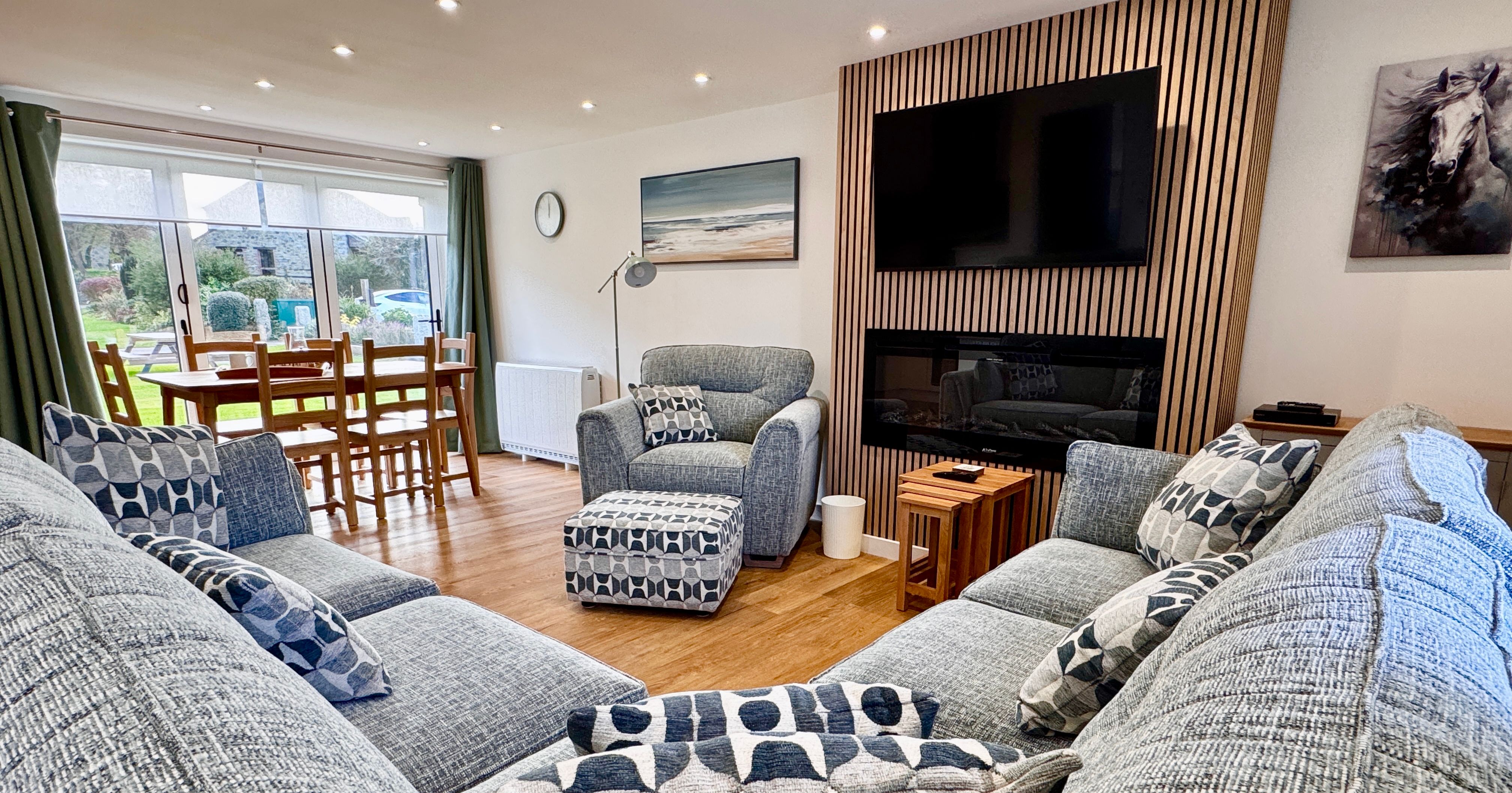 Modern living room with gray sectional sofa and patterned cushions, wooden flooring, wall-mounted TV above a fireplace, dining table by large windows with garden view, and contemporary decor.