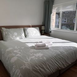 Bright and cozy bedroom with a large bed, floral bedding, two bedside tables with lamps, and sunlight streaming through a window with a view of a stone building outside.