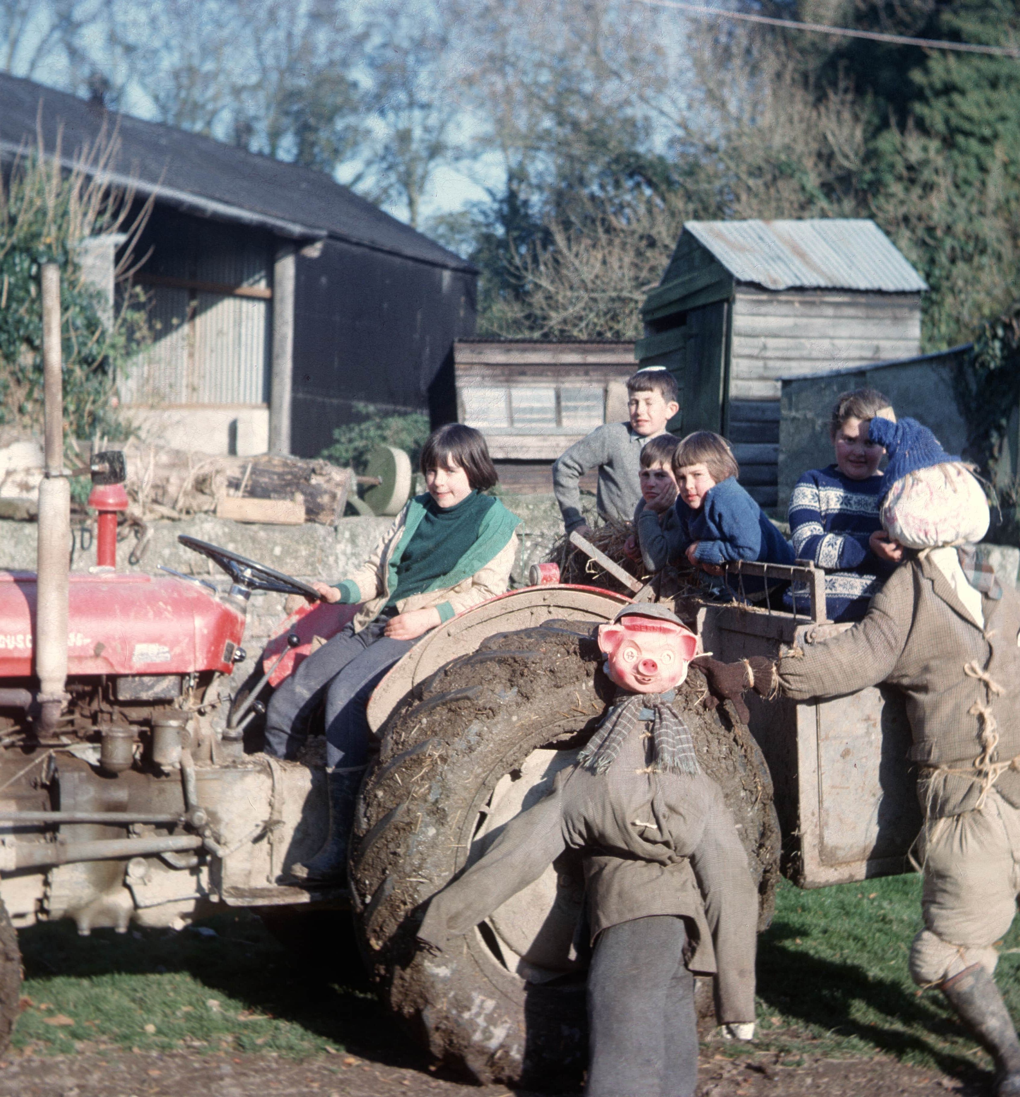 Children sitting on a red Massey Ferguson tractor with two scarecrows, on a farmyard with sheds in the background.