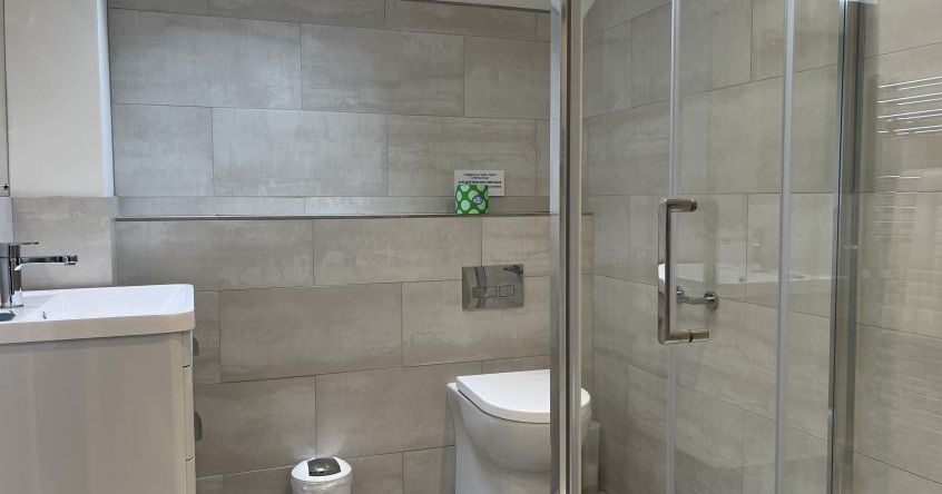 Modern bathroom with light gray tiles, a shower with glass doors, a wall-mounted toilet, a small white trash bin, and a sink with a cabinet.