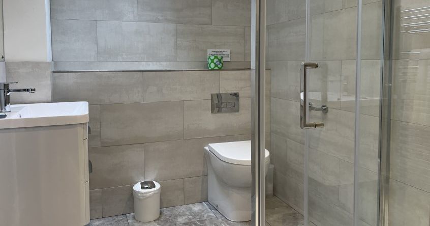 Modern bathroom with light gray tiles, a shower with glass doors, a wall-mounted toilet, a small white trash bin, and a sink with a cabinet.
