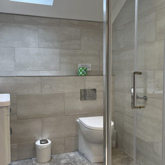 Modern bathroom with light gray tiles, a shower with glass doors, a wall-mounted toilet, a small white trash bin, and a sink with a cabinet.