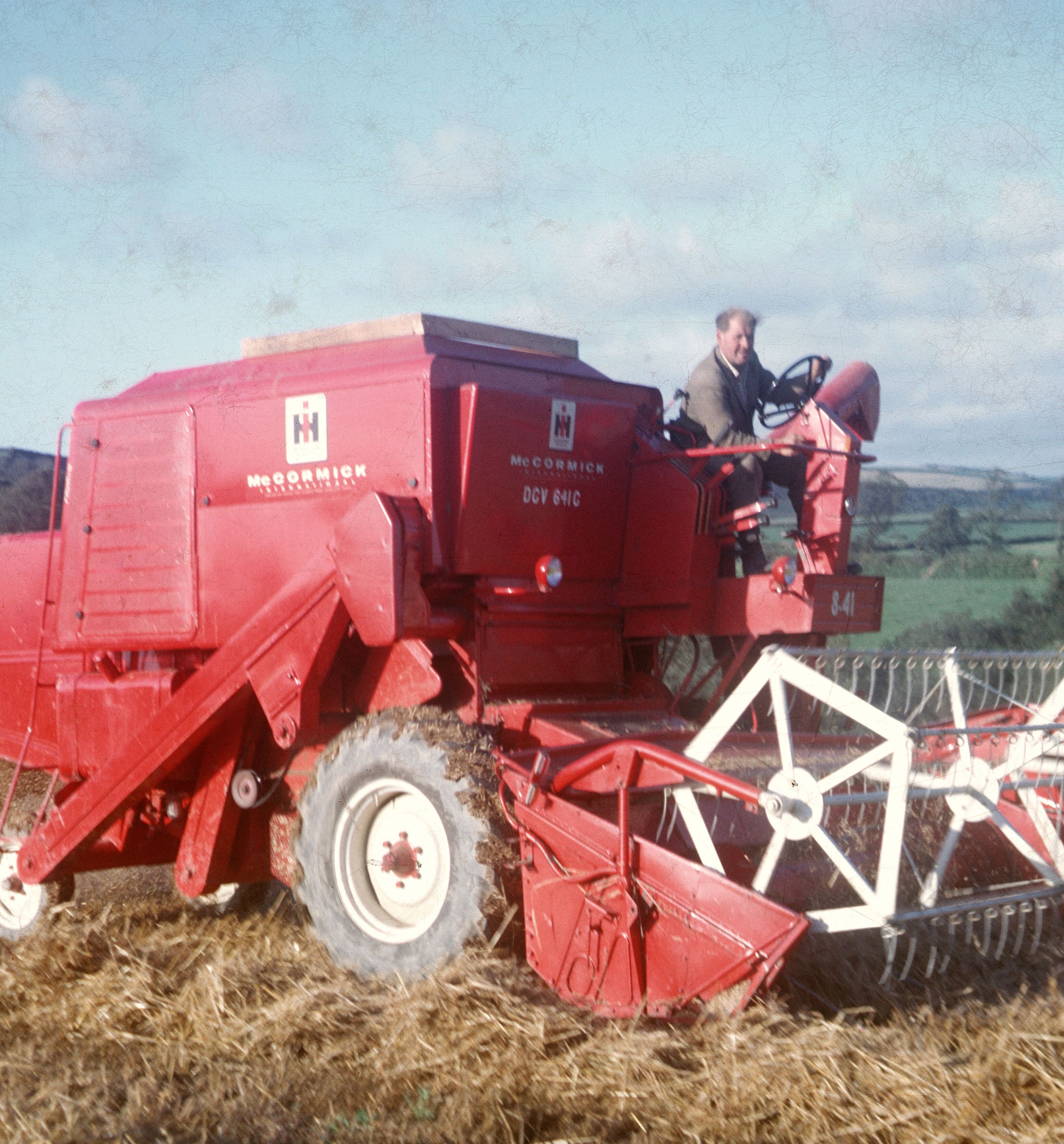 A person operating a red McCormick International 841 combine harvester in a field with hay on the ground and green countryside in the background.