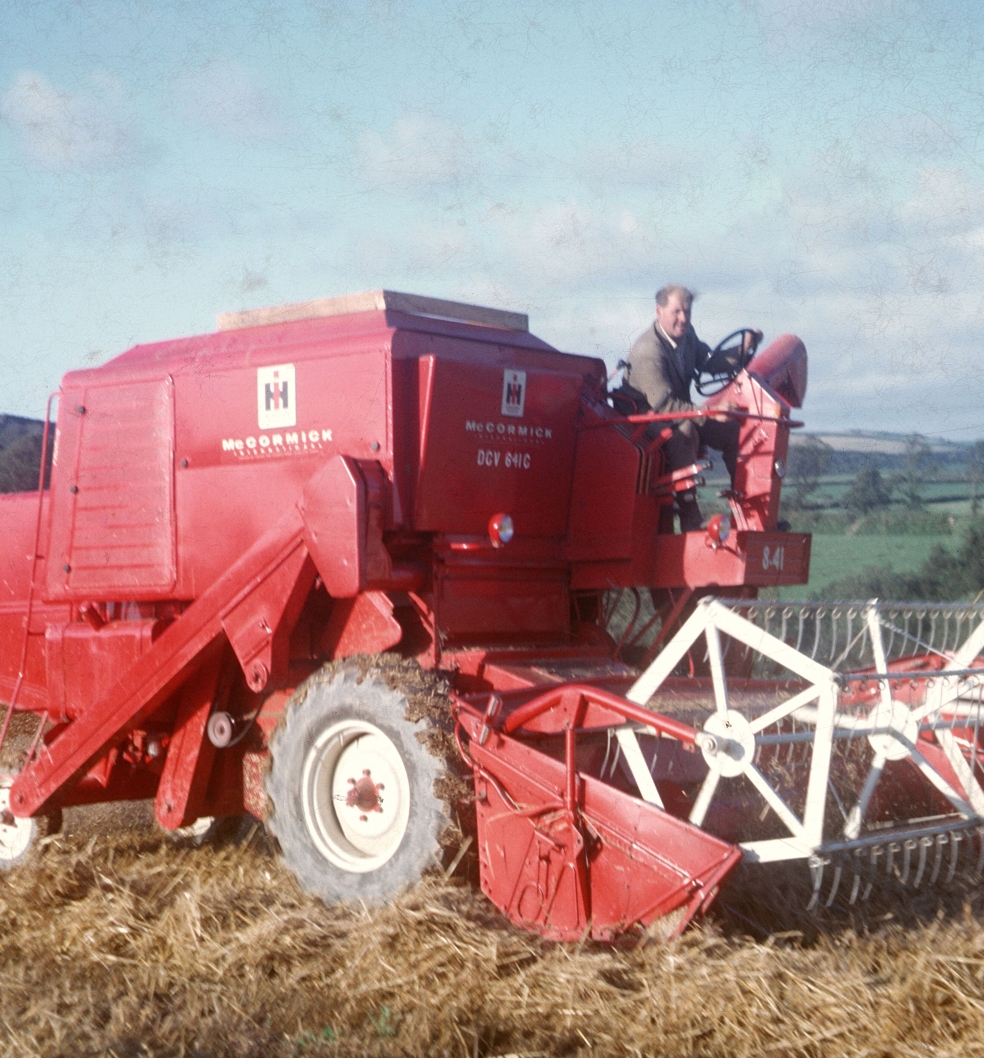 A person operating a red McCormick International 841 combine harvester in a field with hay on the ground and green countryside in the background.