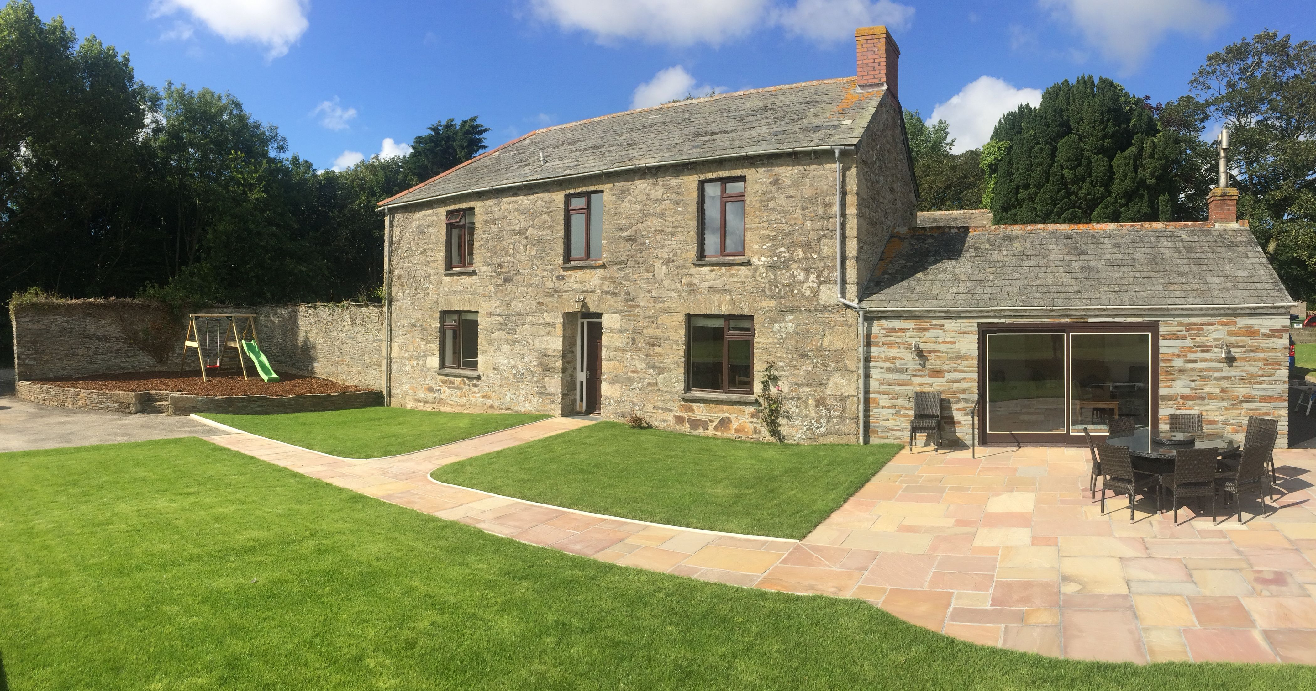 A stone farmhouse with a large tiled patio, green lawn, outdoor dining set, and a swing set in the yard. Surrounded by trees under a bright blue sky with scattered clouds.