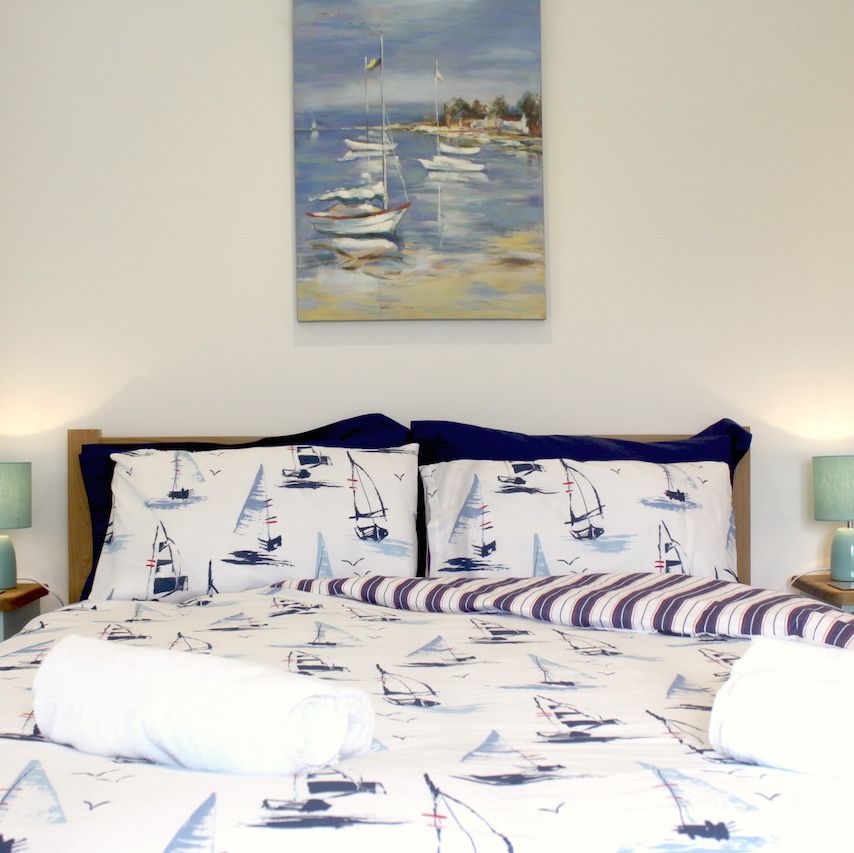A neatly made bed with sailboat-themed bedding, flanked by two bedside tables with matching turquoise lamps, and a seascape painting hanging above the headboard.