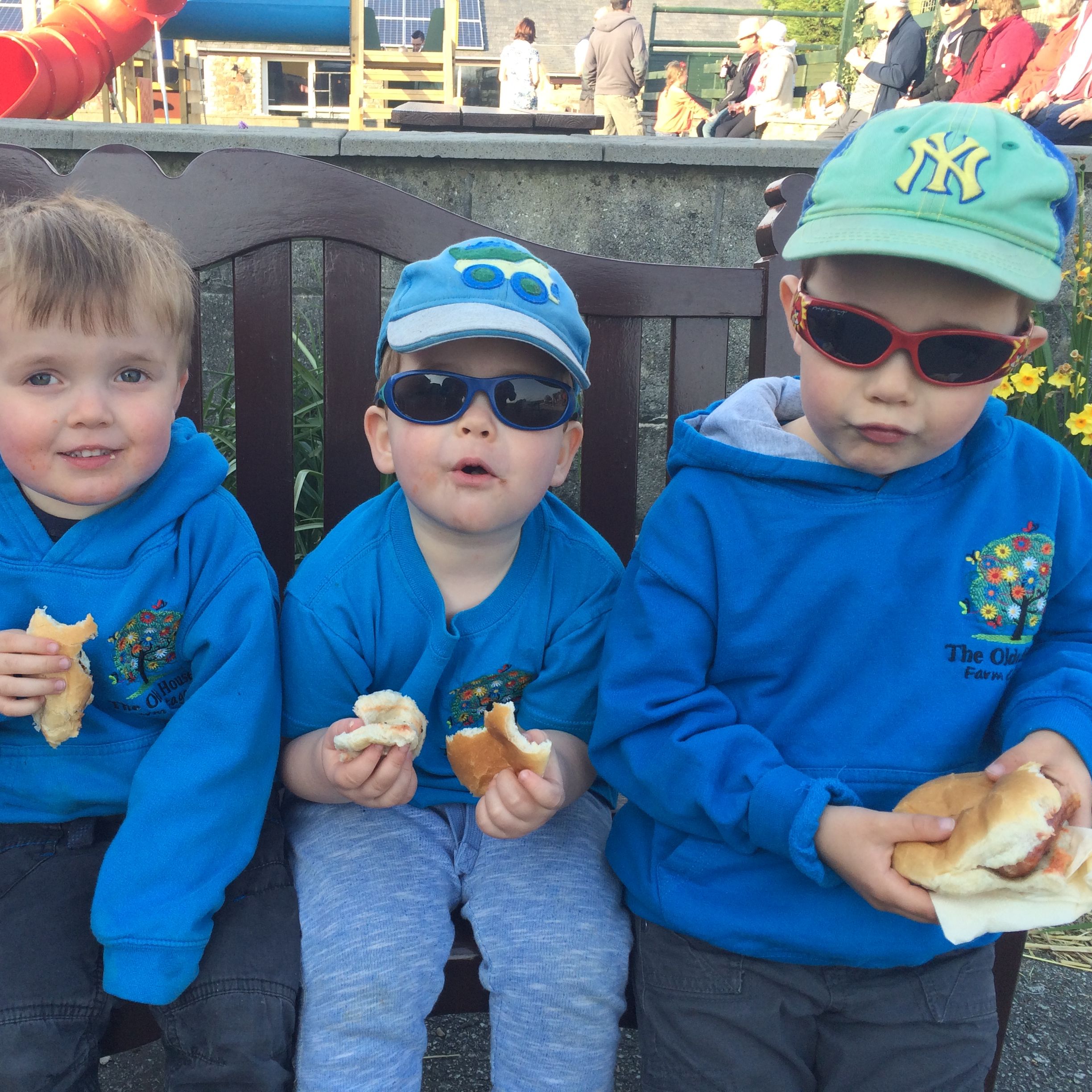 Three young children sitting on a bench, wearing blue clothes and sunglasses, eating sandwiches or burgers.