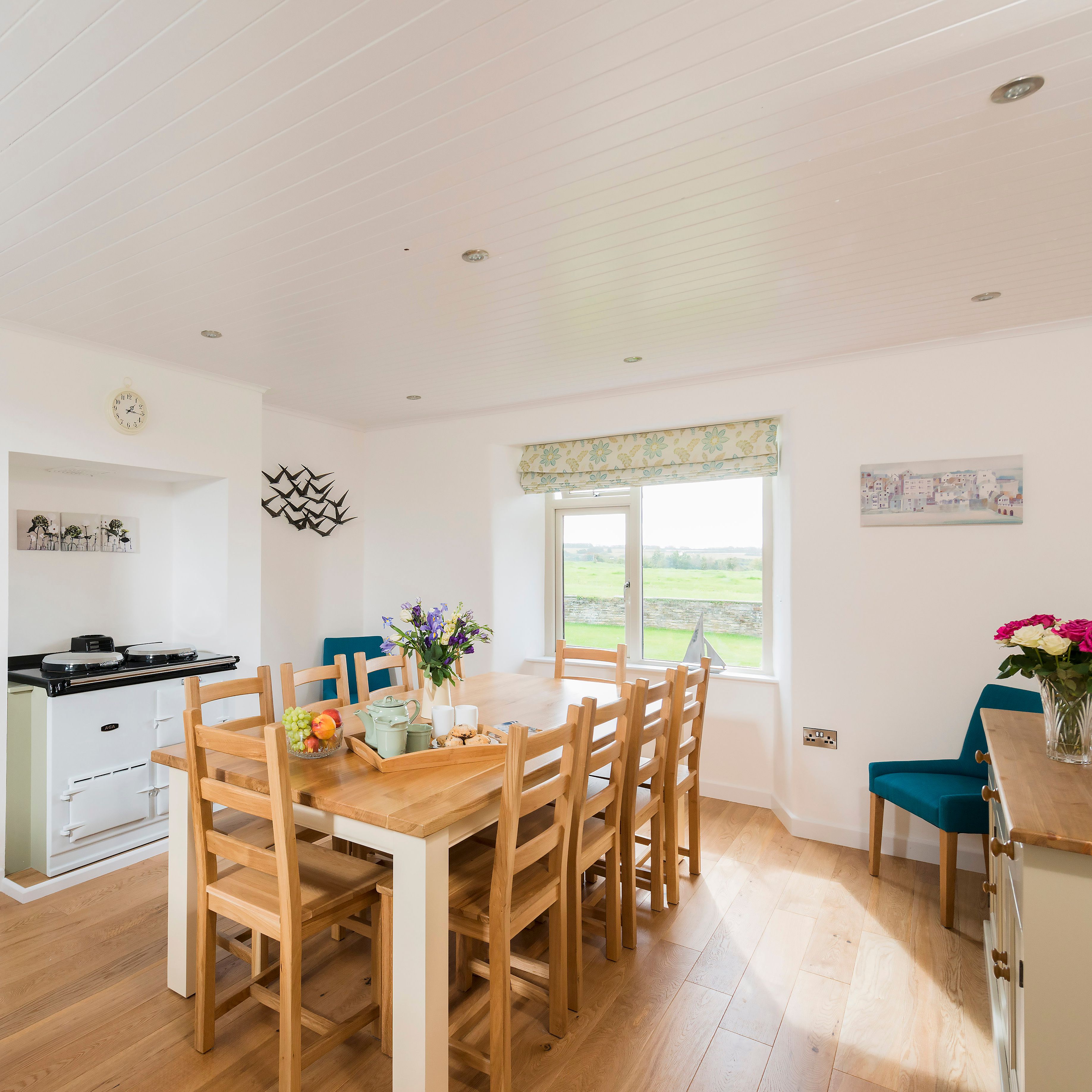 Bright and spacious dining room with a wooden table and chairs, fresh flowers, light wood flooring, large window with scenic countryside views, and adjoining living room with fireplace.