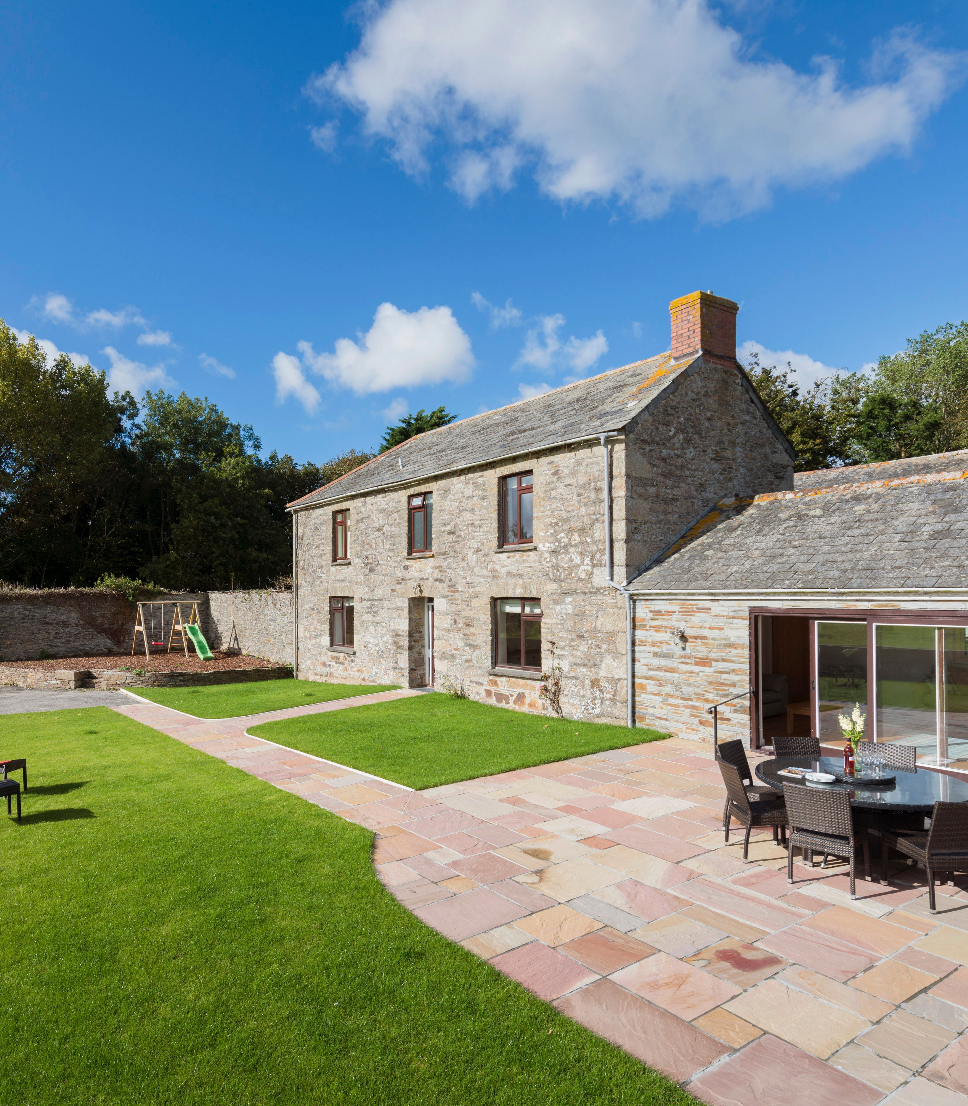 Stone farmhouse with patio and outdoor dining set, surrounded by green lawn, trees, and a children's playset under a blue sky with scattered clouds.