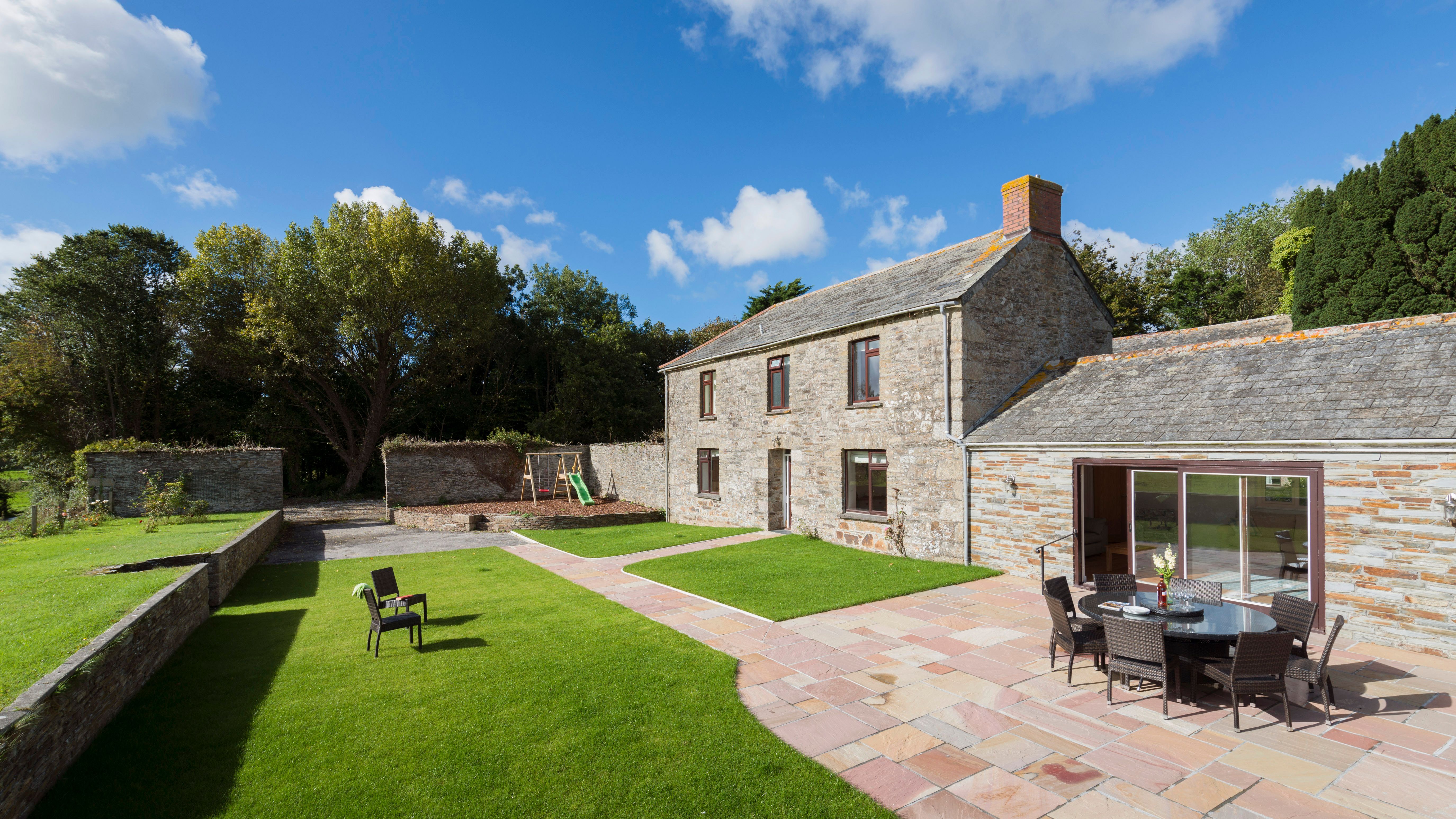 Stone farmhouse with patio and outdoor dining set, surrounded by green lawn, trees, and a children's playset under a blue sky with scattered clouds.